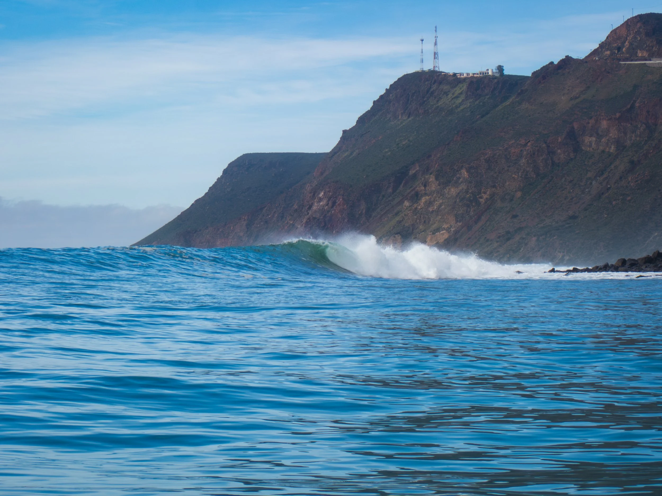 Ocean waves near a rocky cliff with communication towers and buildings on top.