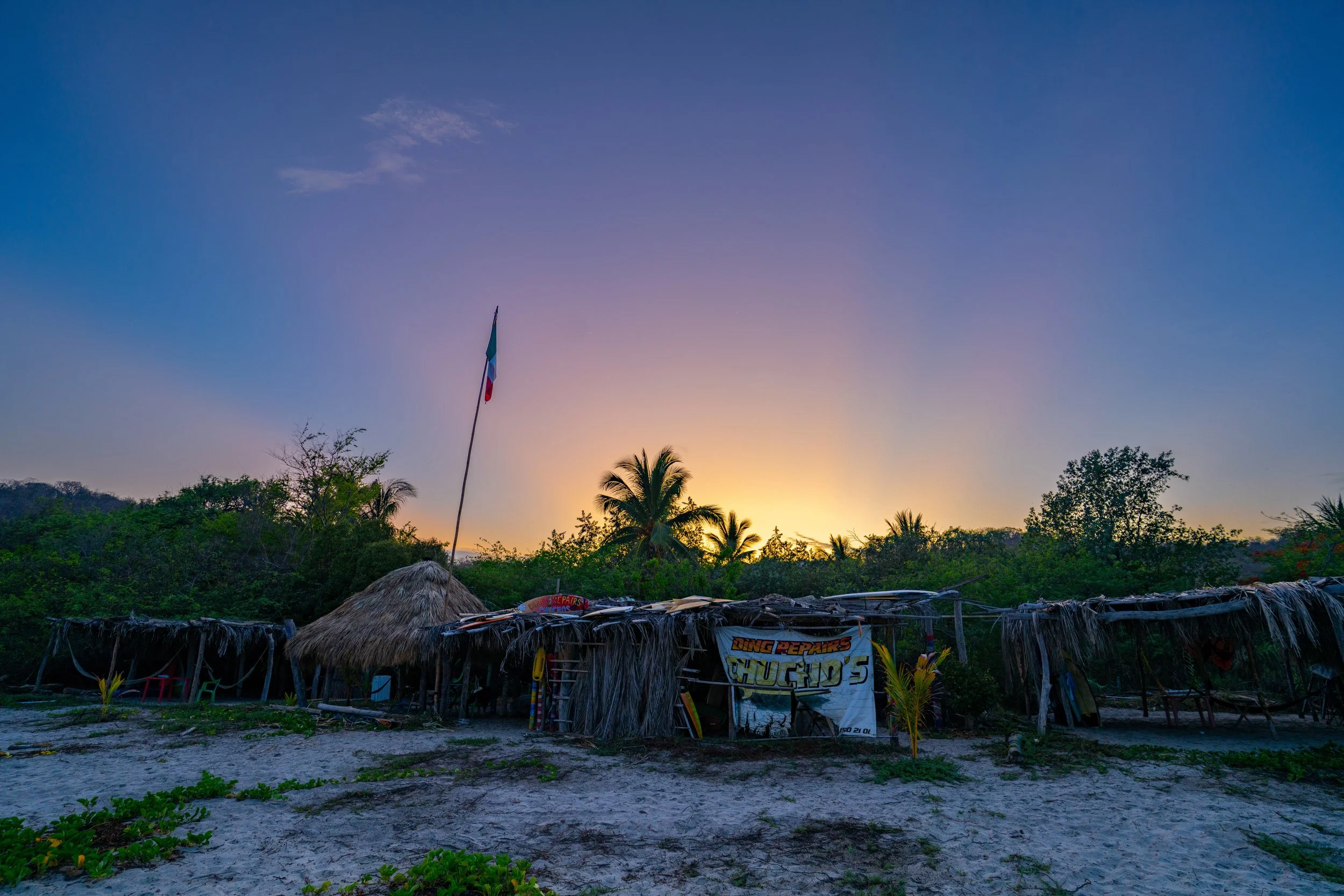 Sunset over tropical trees and a thatched-roof hut with a flag and colorful signs in the foreground.