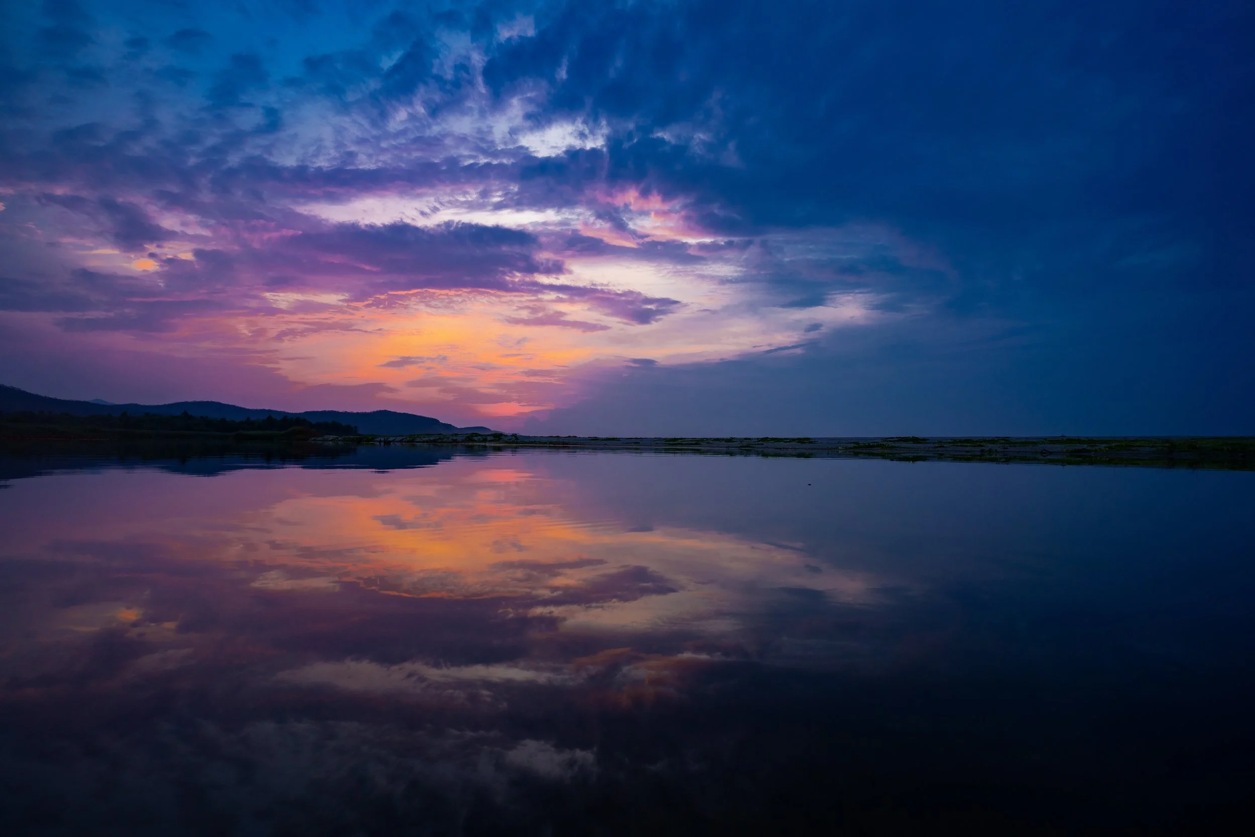 A sunset over a body of water with colorful clouds and distant hills, reflected on the surface of the water.