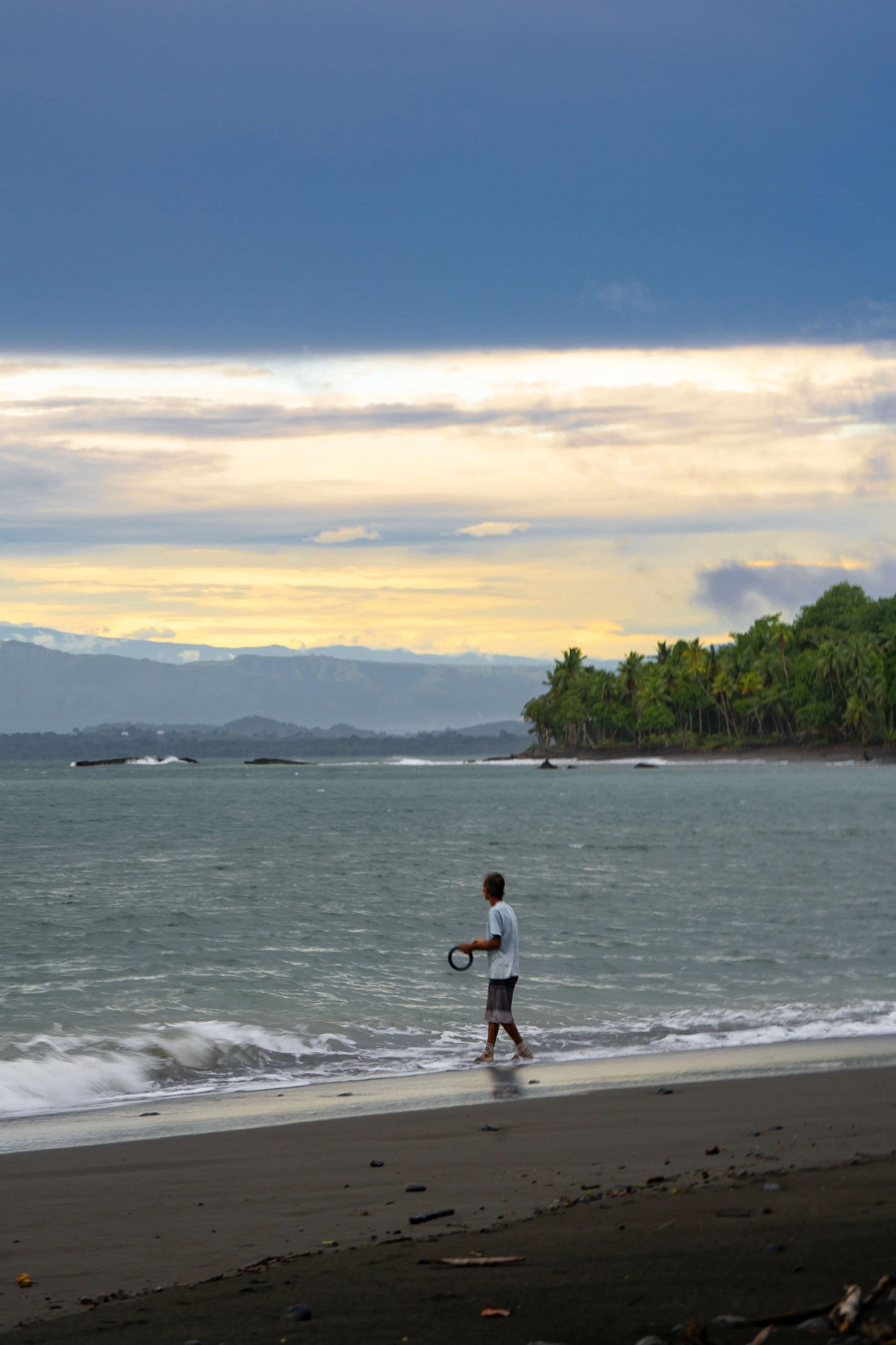 A person walking along the beach with mountains, water, and a cloudy sky in the background during sunset.