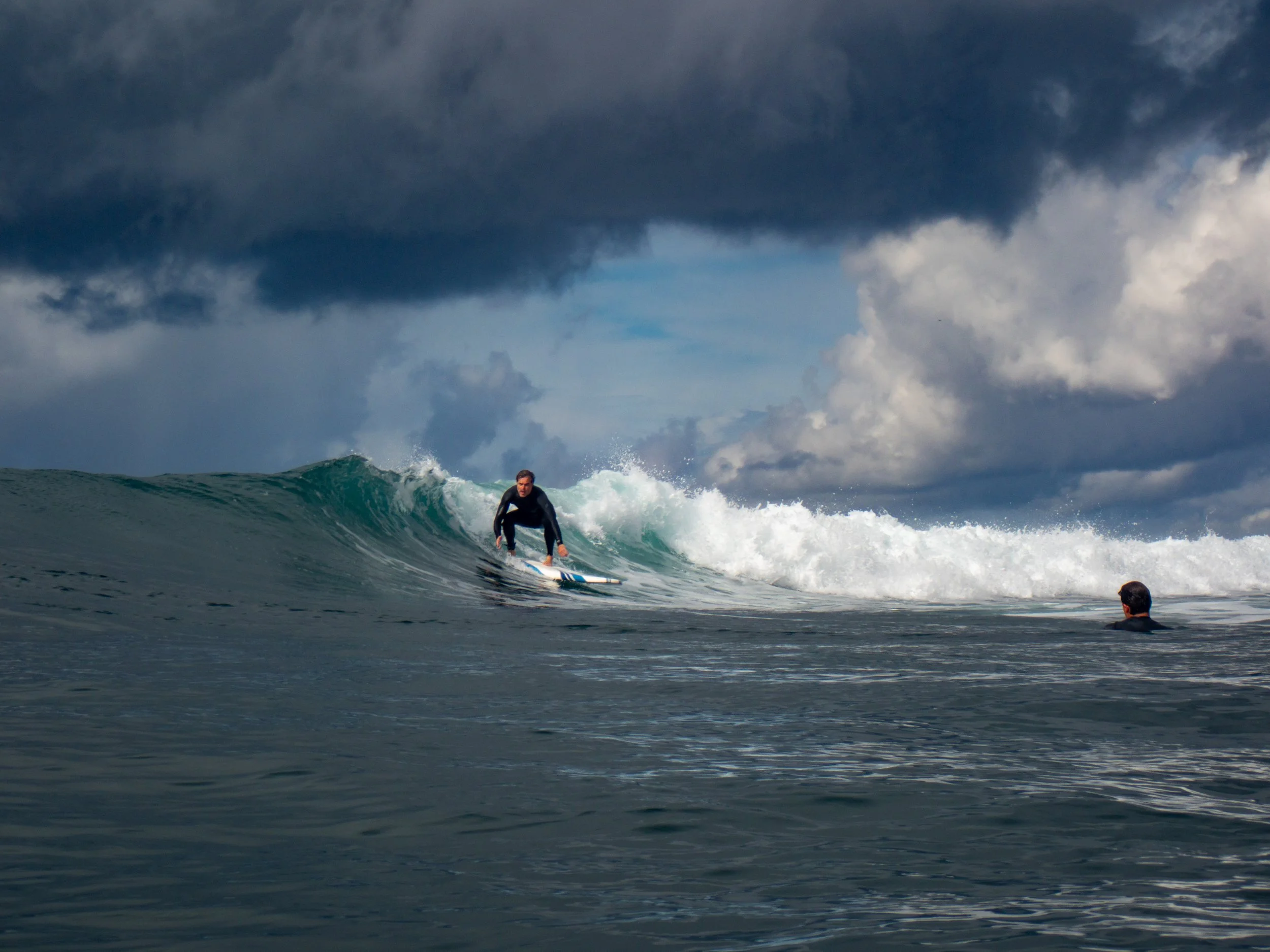 A person surfing on a wave in the ocean with dark stormy clouds overhead and another person in the water watching.