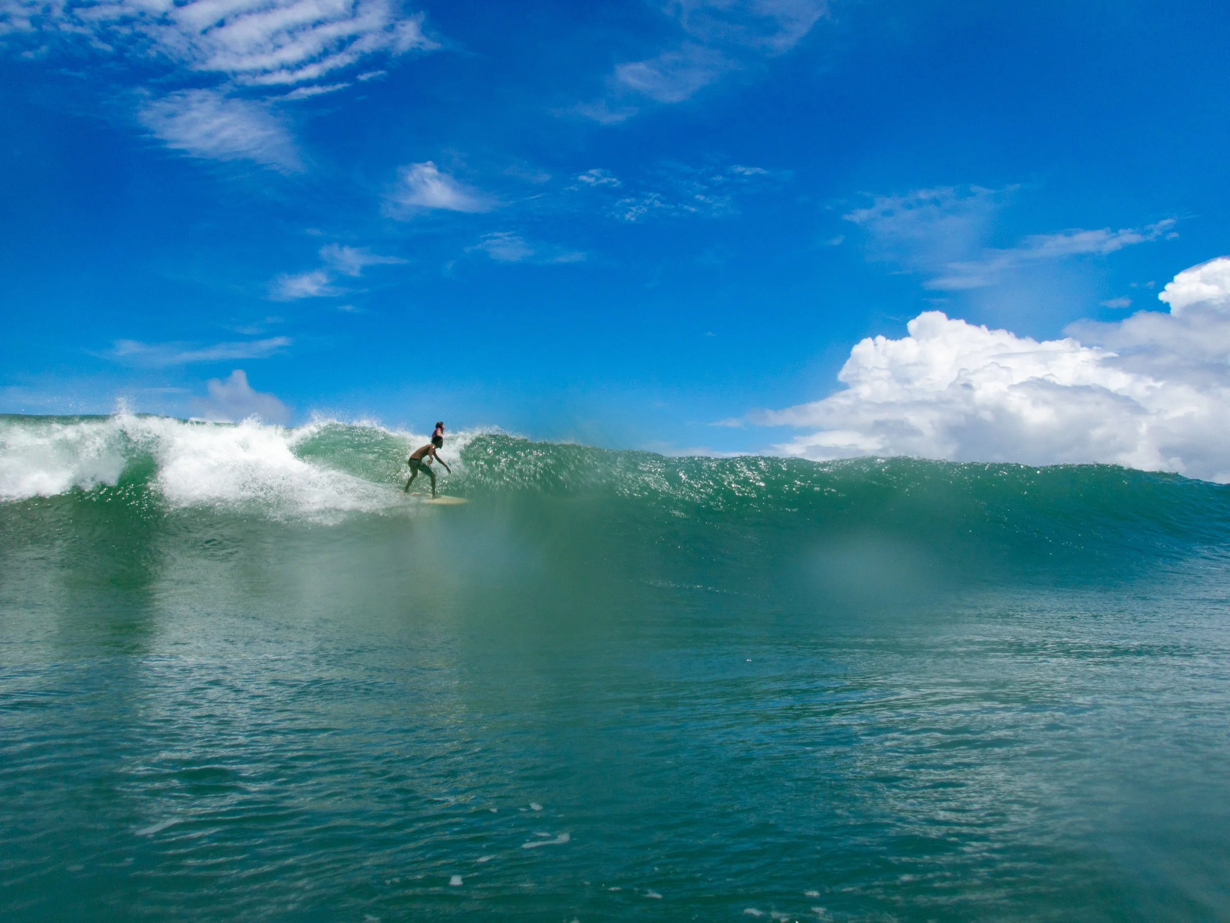 Surfer riding a wave with a clear blue sky and scattered clouds in the background.