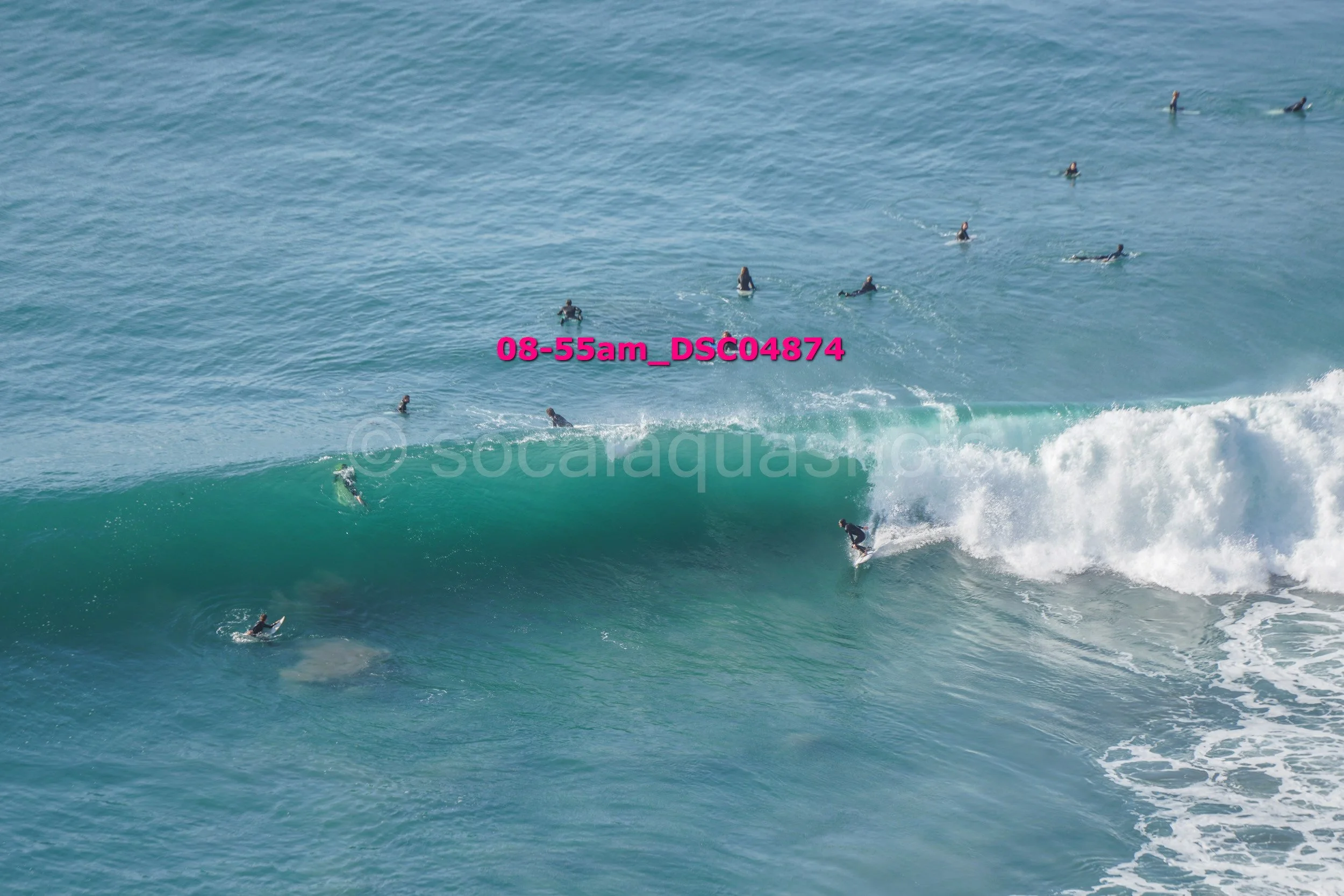 Surfers riding a large ocean wave with others waiting in the water, during daytime.