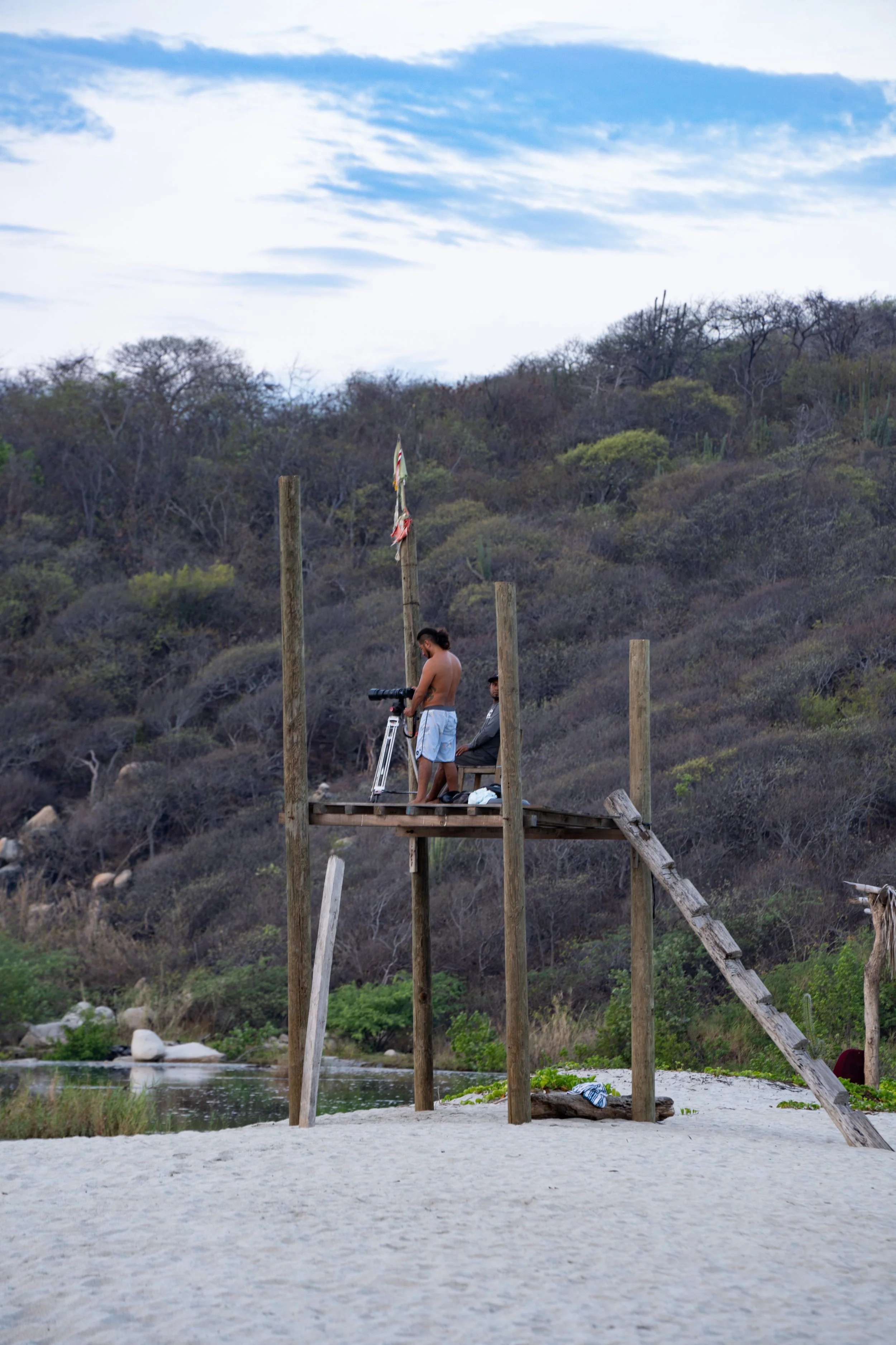 Two men building a wooden structure on a sandy beach next to a river or lake, with mountains and shrubs in the background.