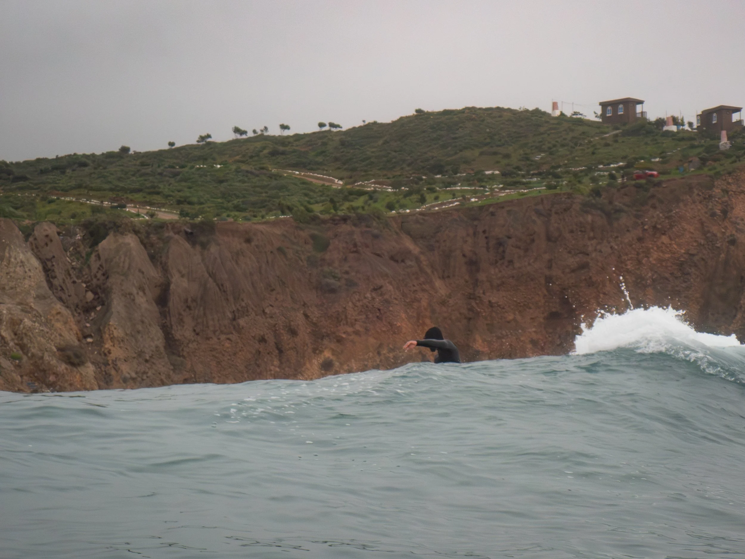 Person in a wetsuit surfing near rocky cliffs with residential houses on a green hillside under an overcast sky.