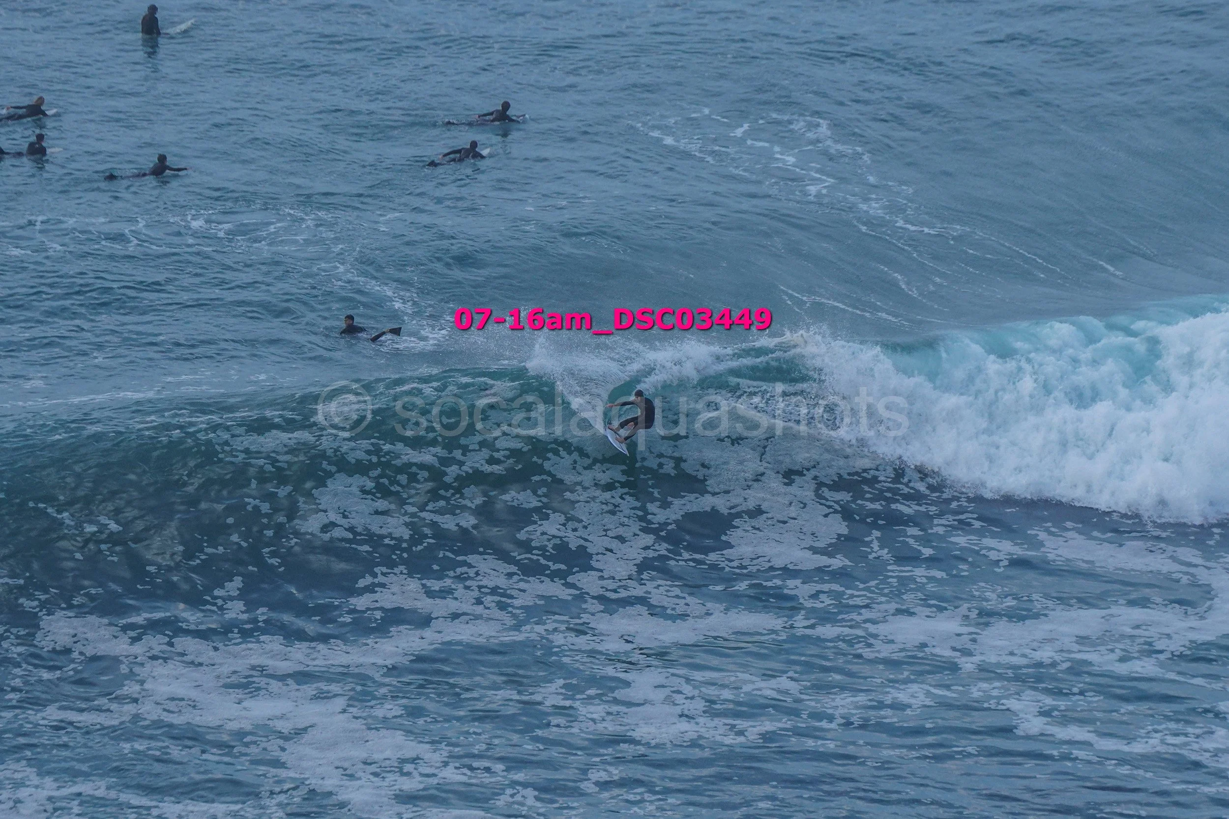 Multiple surfers in the ocean, with one surfing a wave in the foreground and others floating or swimming in the background.