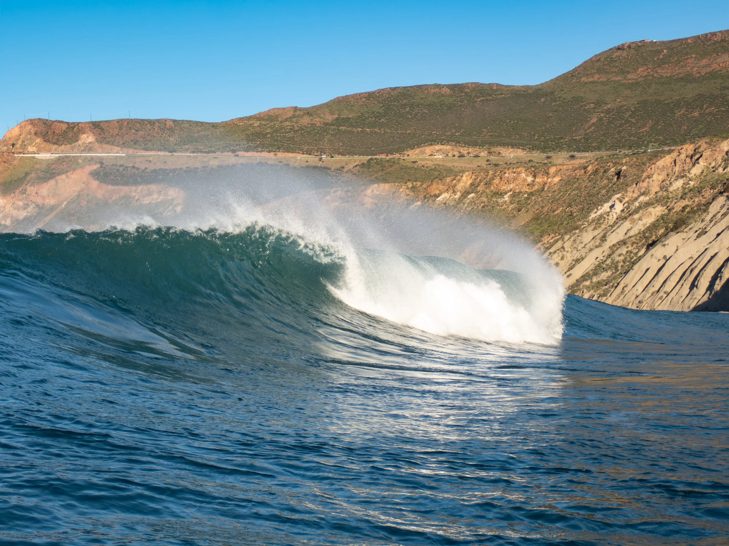 A large ocean wave curling near rocky cliffs with green hills in the background on a clear sunny day.