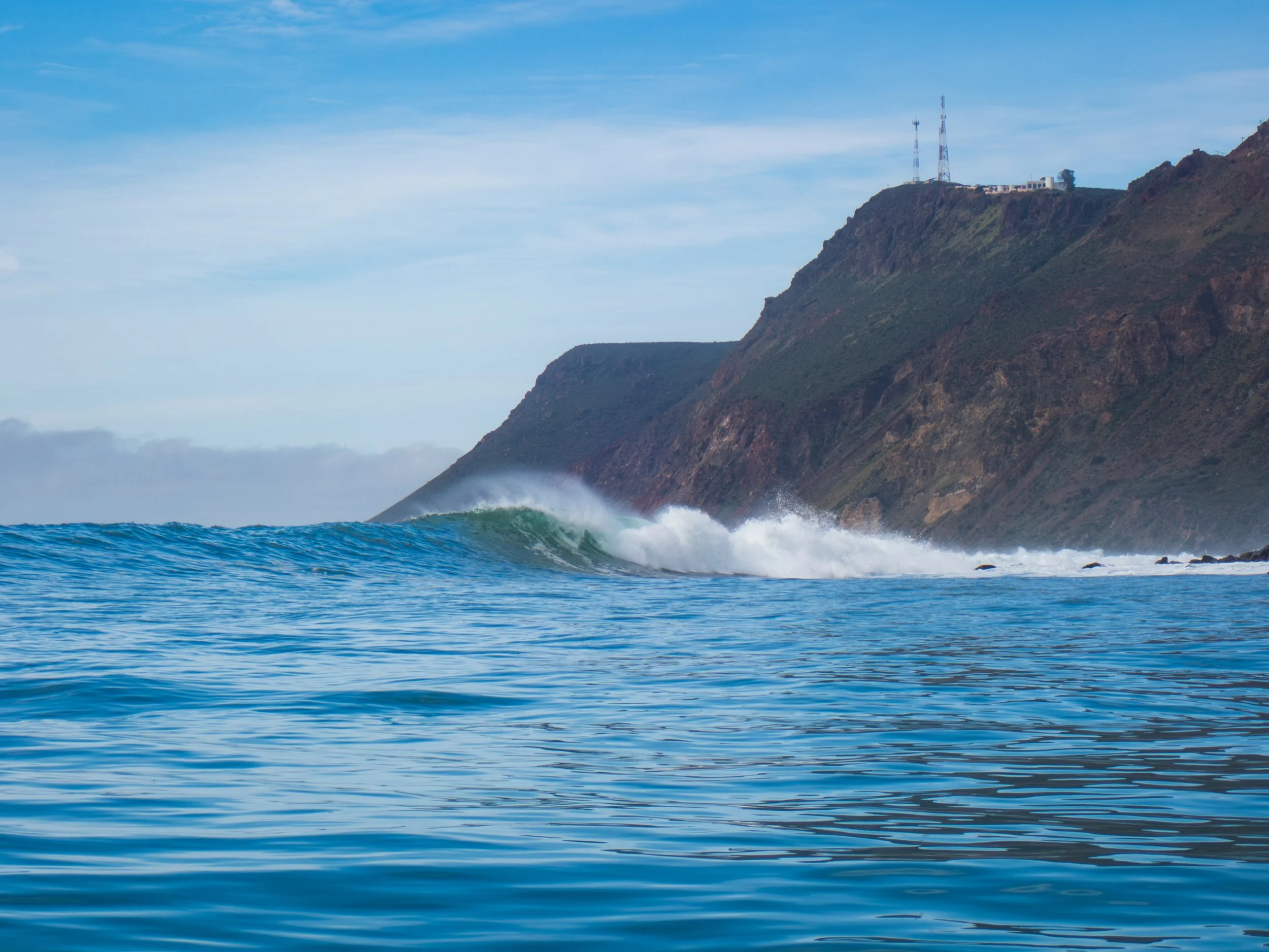 Ocean waves with a hill in the background that has communication towers on top.