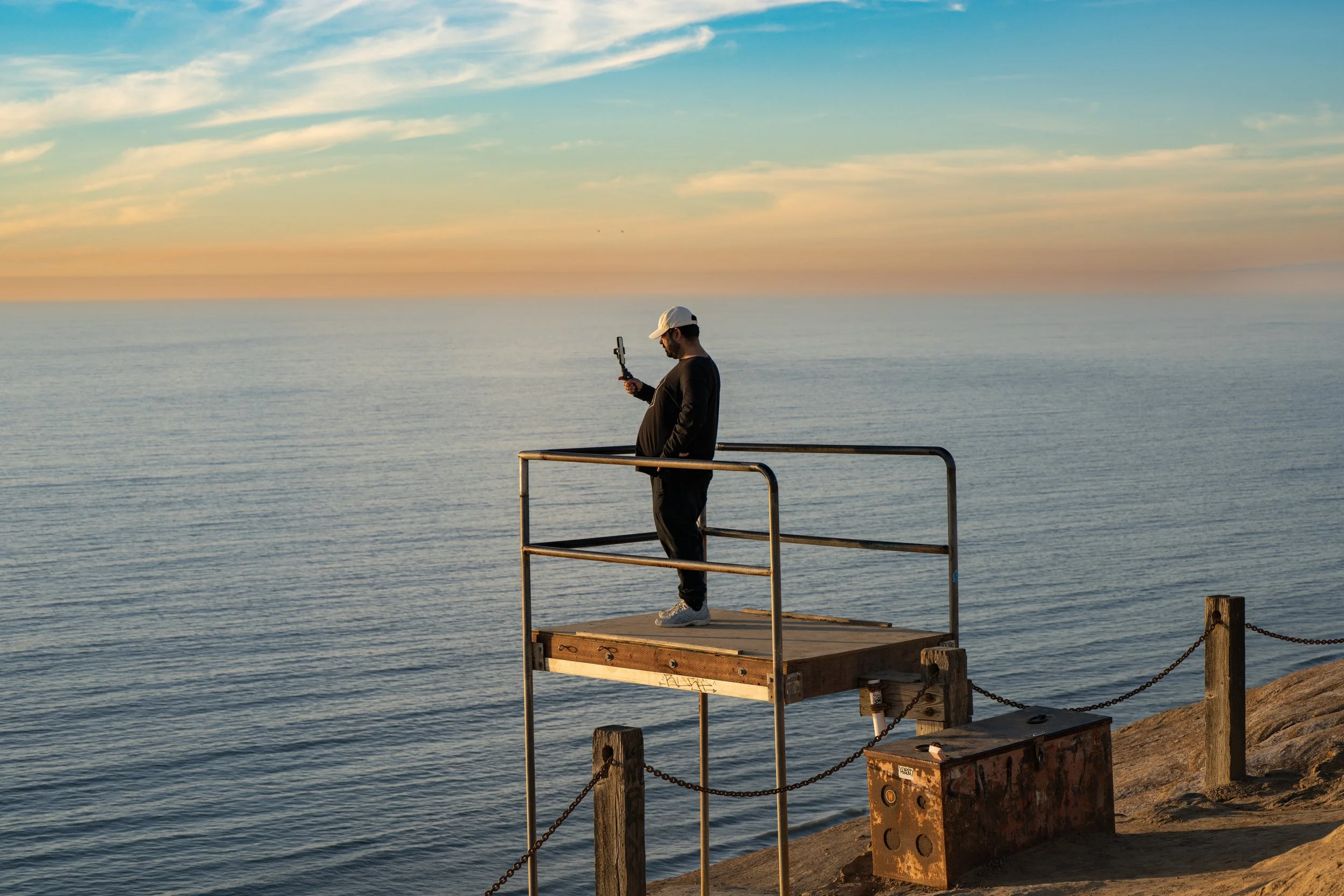 A man standing on a wooden platform over the water, using a selfie stick, overlooking the calm ocean at sunset.