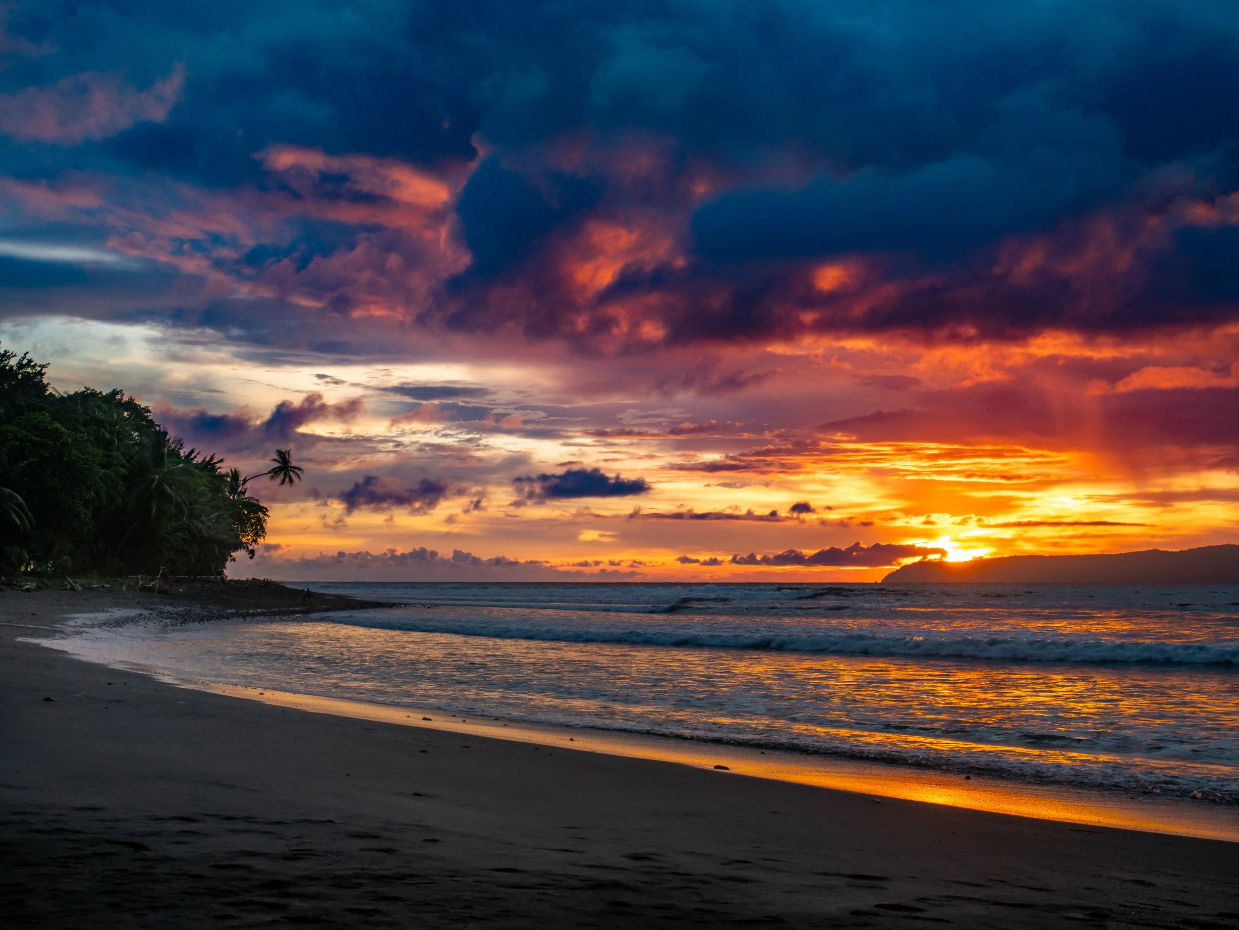 Sunset over a beach with palm trees, colorful sky, and ocean waves.