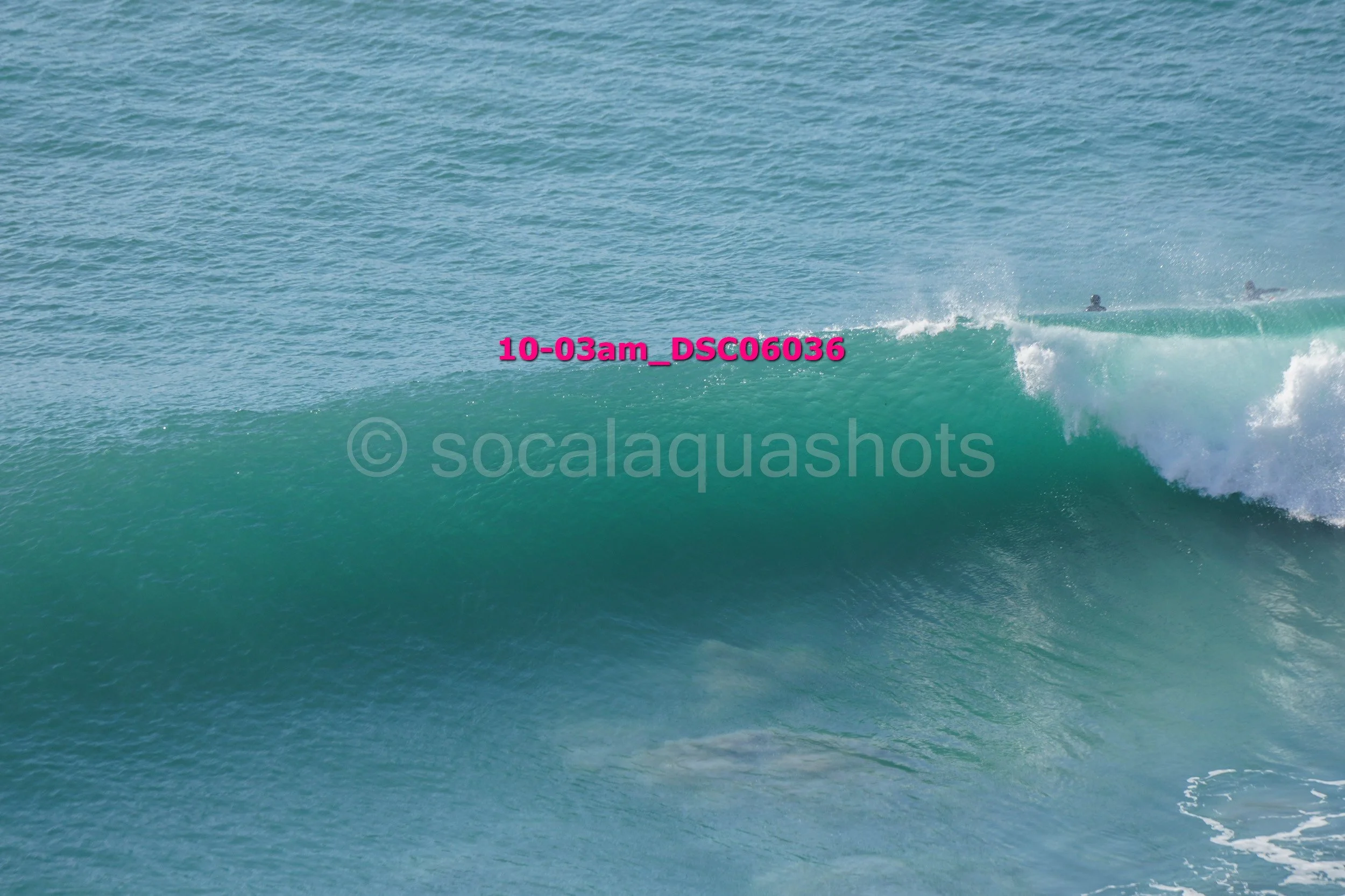 Large ocean wave with surfers in the distance and a watermark from socalquashots.