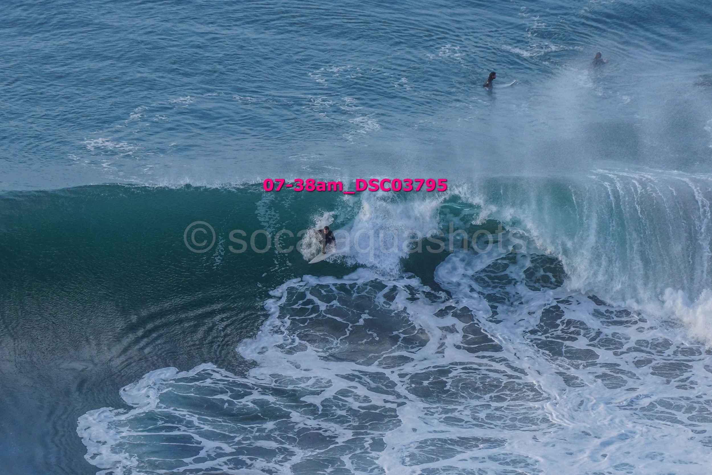 A person surfing on a large ocean wave with three other surfers visible in the background.