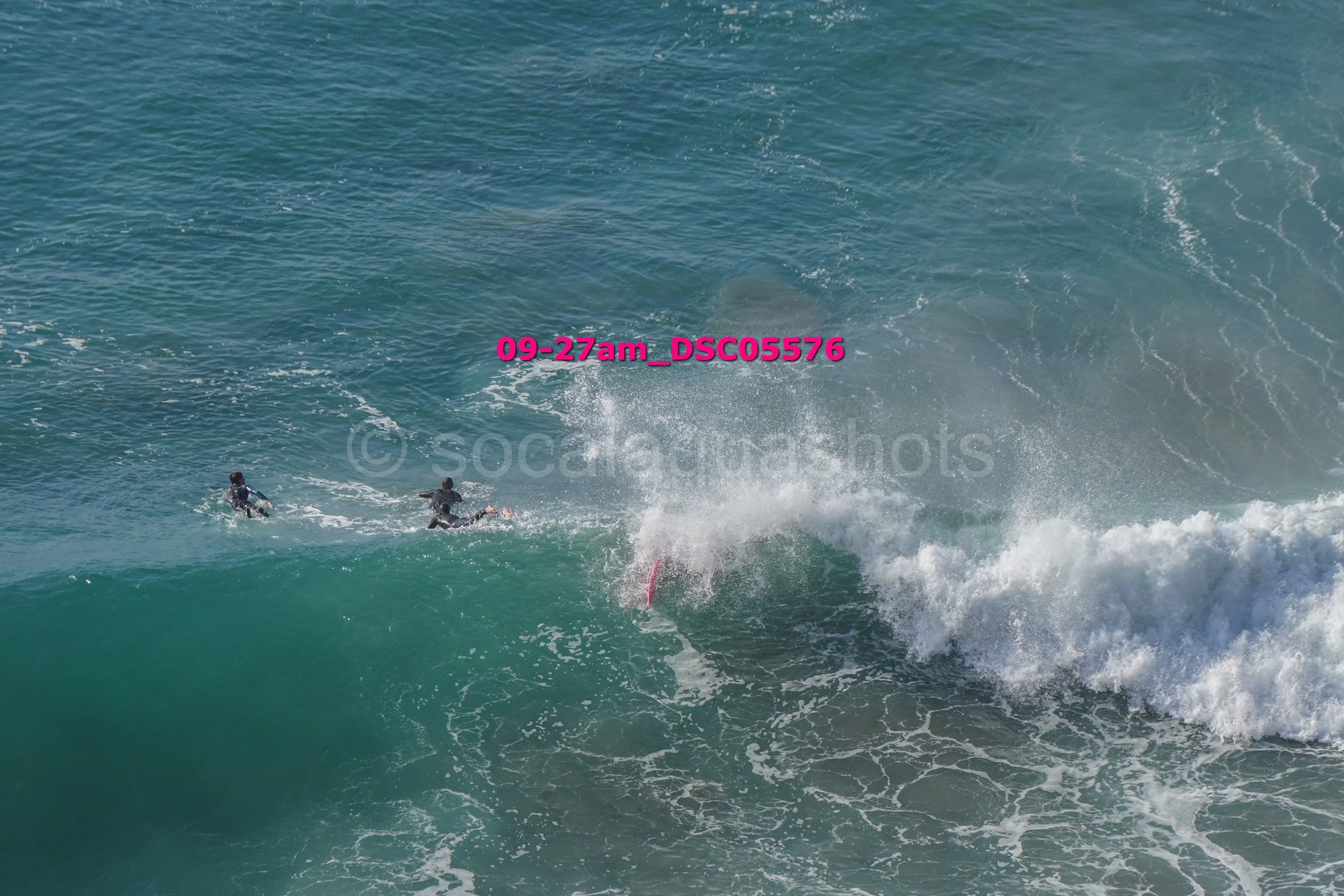 Two surfers in wetsuits riding a large wave at the beach, with spray and foam in the ocean water.