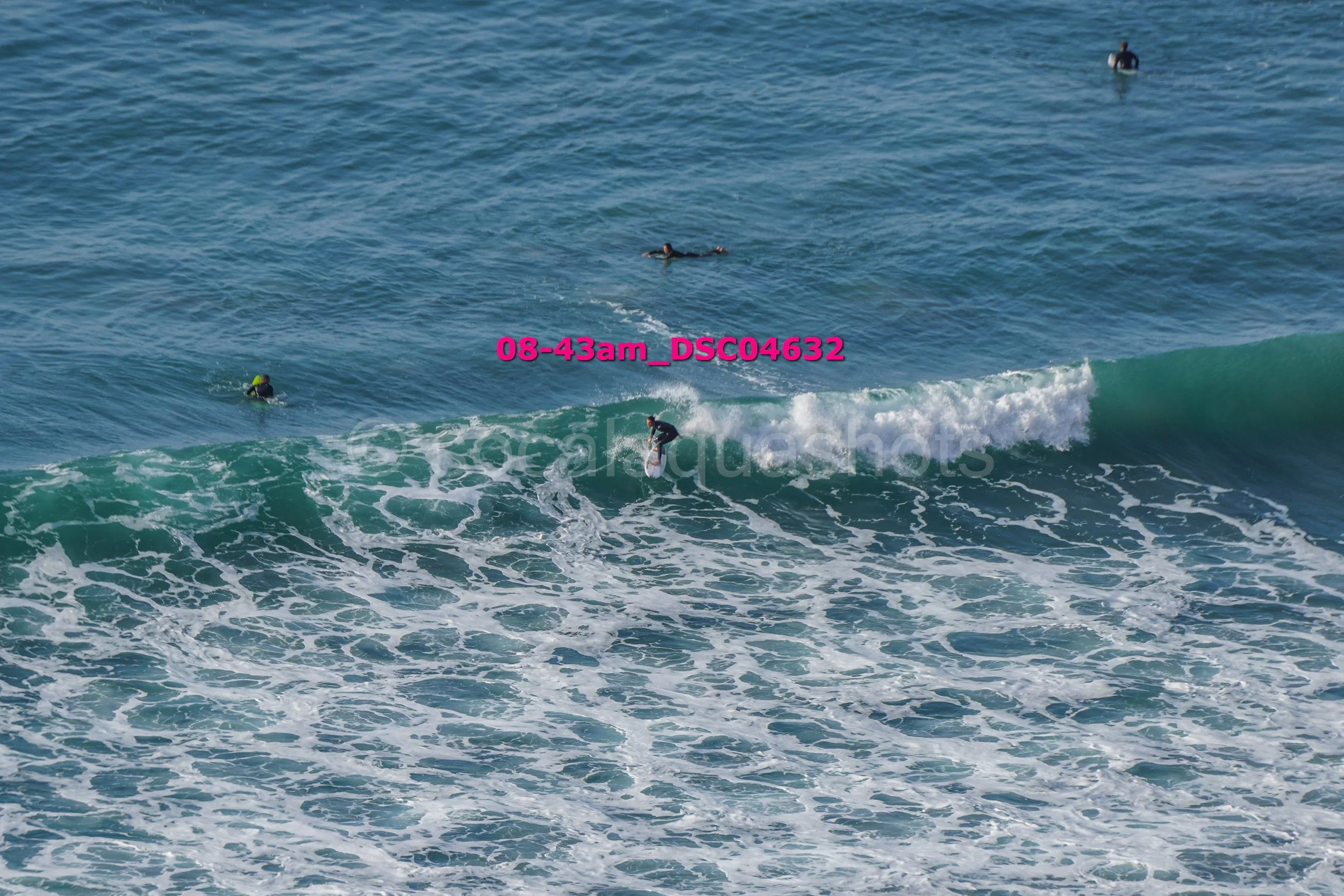 A person surfing on a wave in the ocean, with several people swimming and floating in the water nearby.