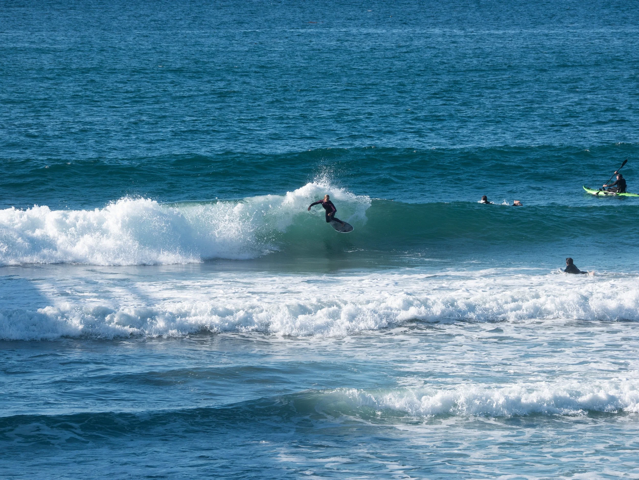 A person surfing on a wave with several others in the water and on a paddleboard nearby.
