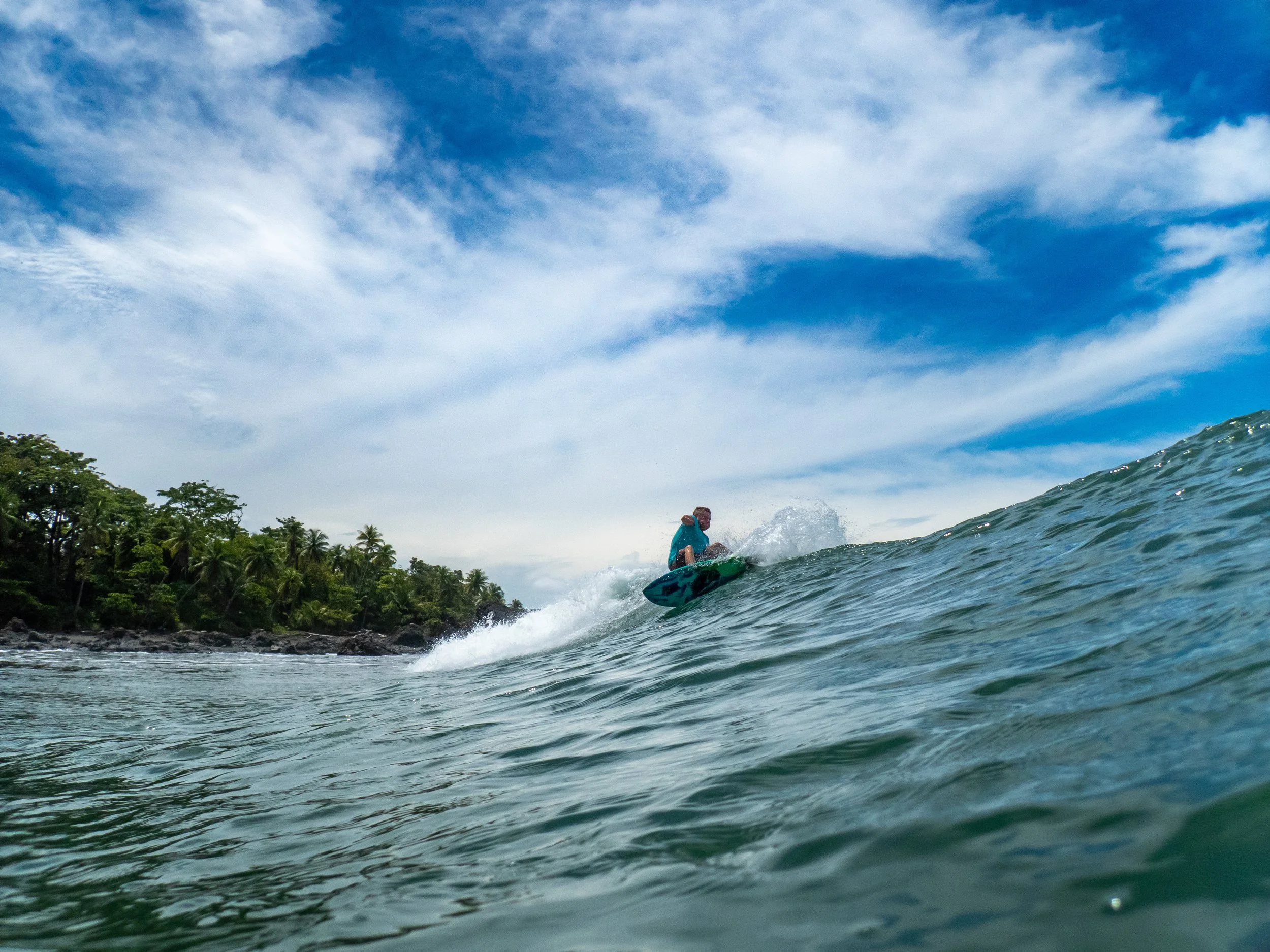 Person surfing on an ocean wave near a tropical island with lush trees and a partly cloudy sky.