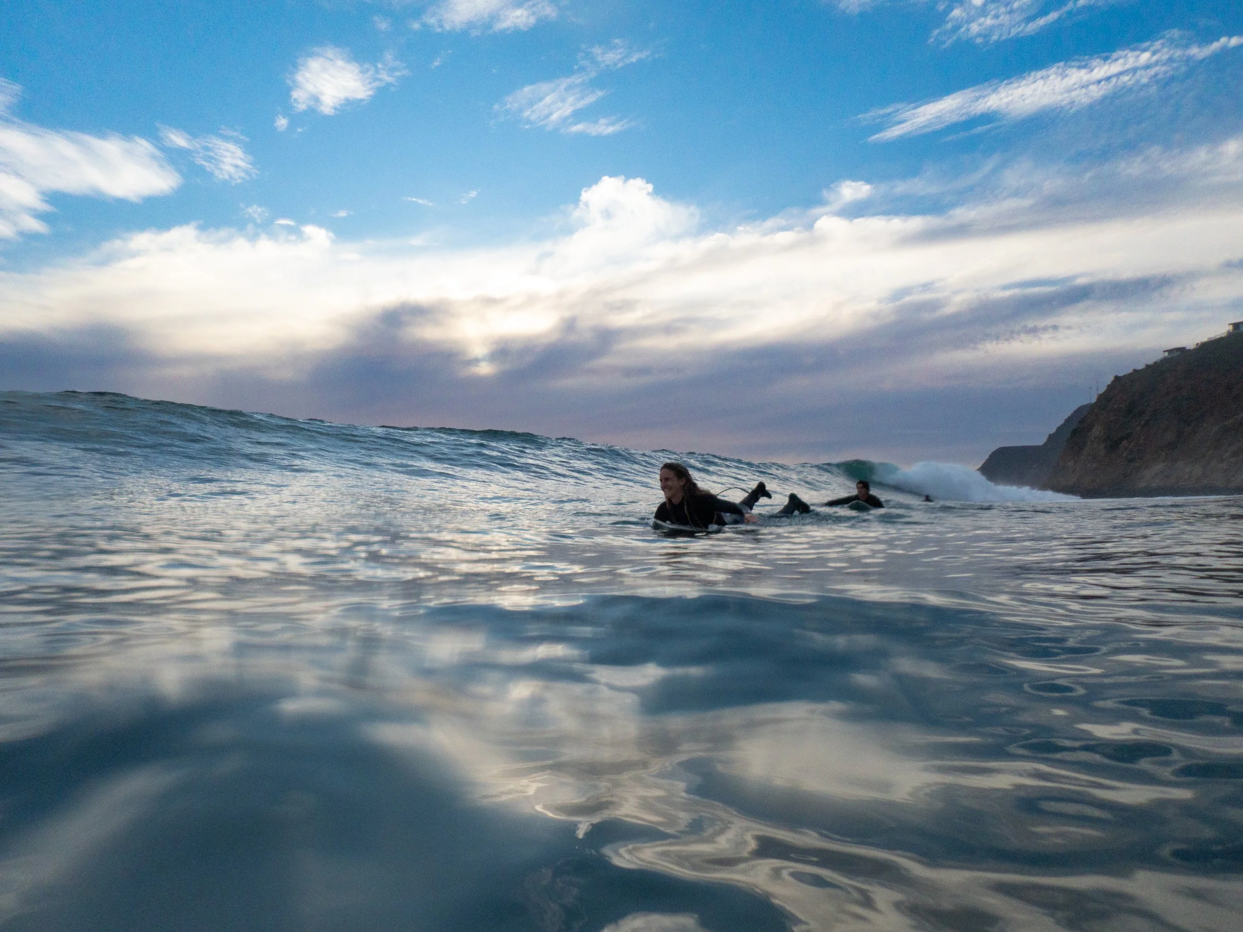 People surfing in the ocean near coastline during sunset, with cloudy sky above.