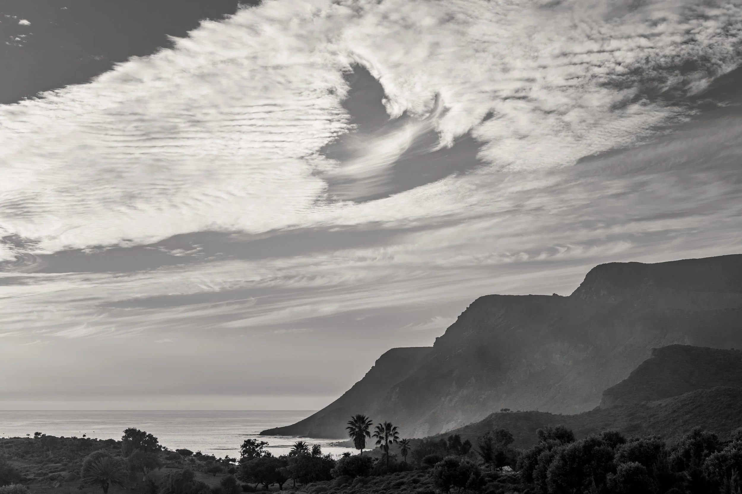 Black and white landscape with large mountain cliffs on the right, a body of water at the base, and a sky filled with clouds above.