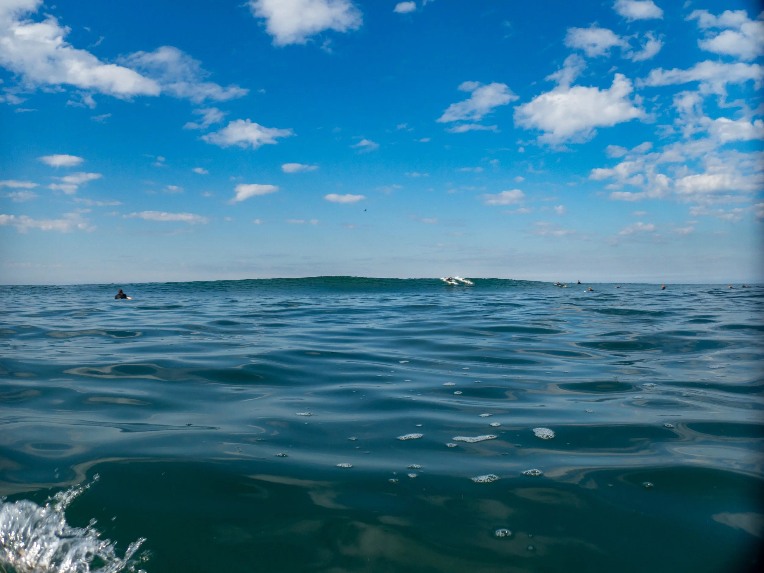 Ocean with a clear blue sky and scattered clouds, several surfers in the water, some riding small waves.