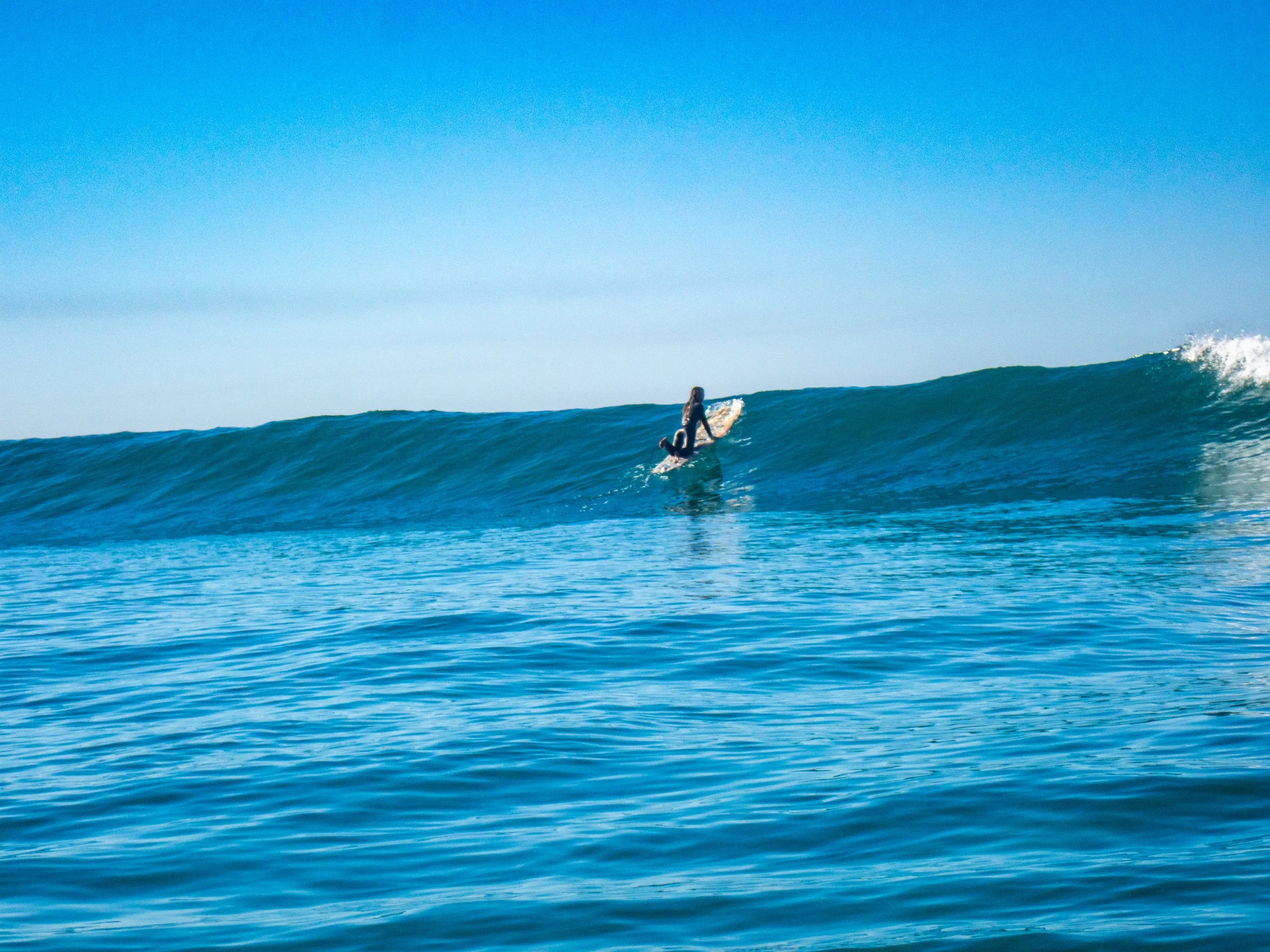A person riding a surfboard on a large blue wave in the ocean under a clear blue sky.
