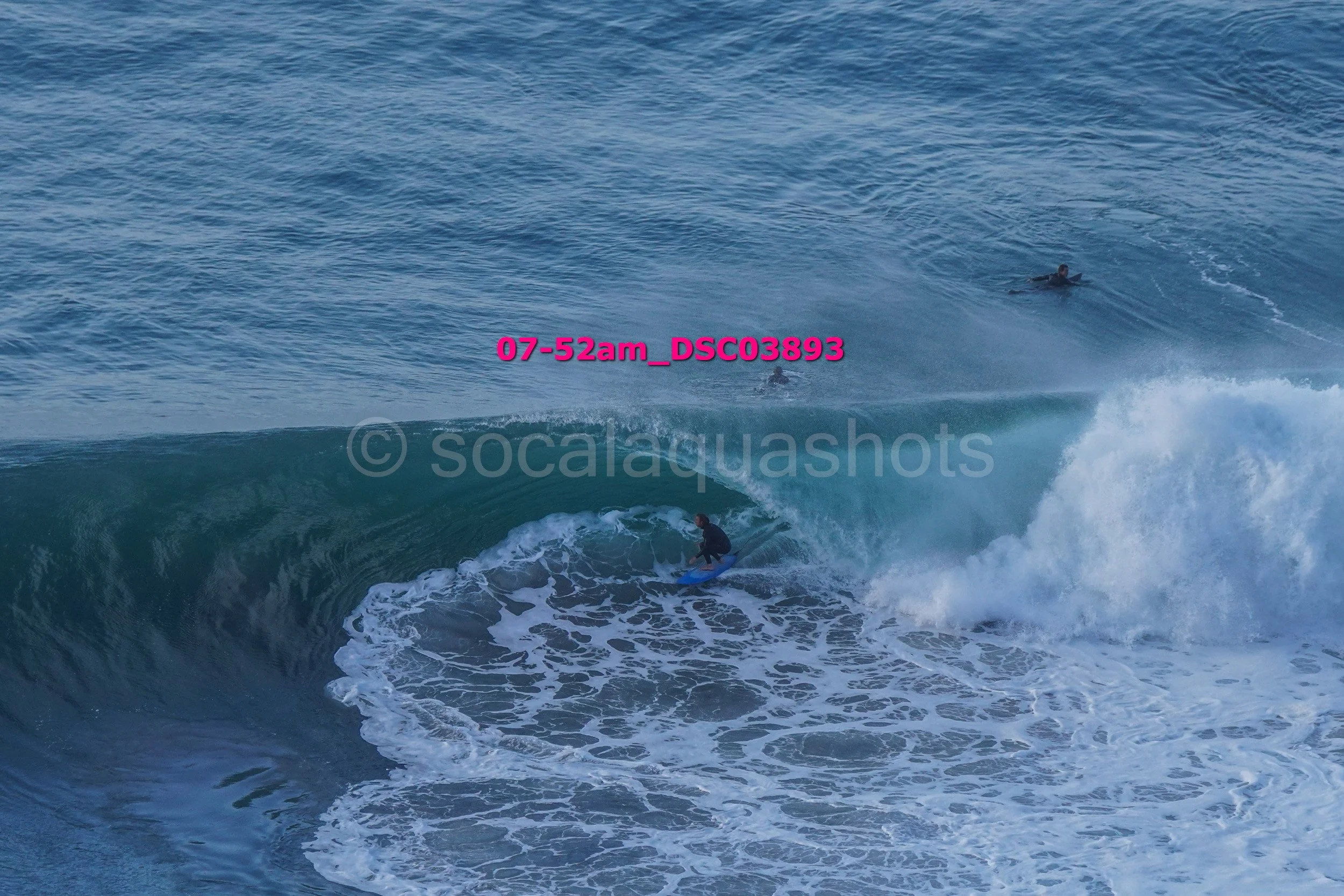 A surfer rides a large ocean wave with two other surfers visible in the background, swimming in the water.