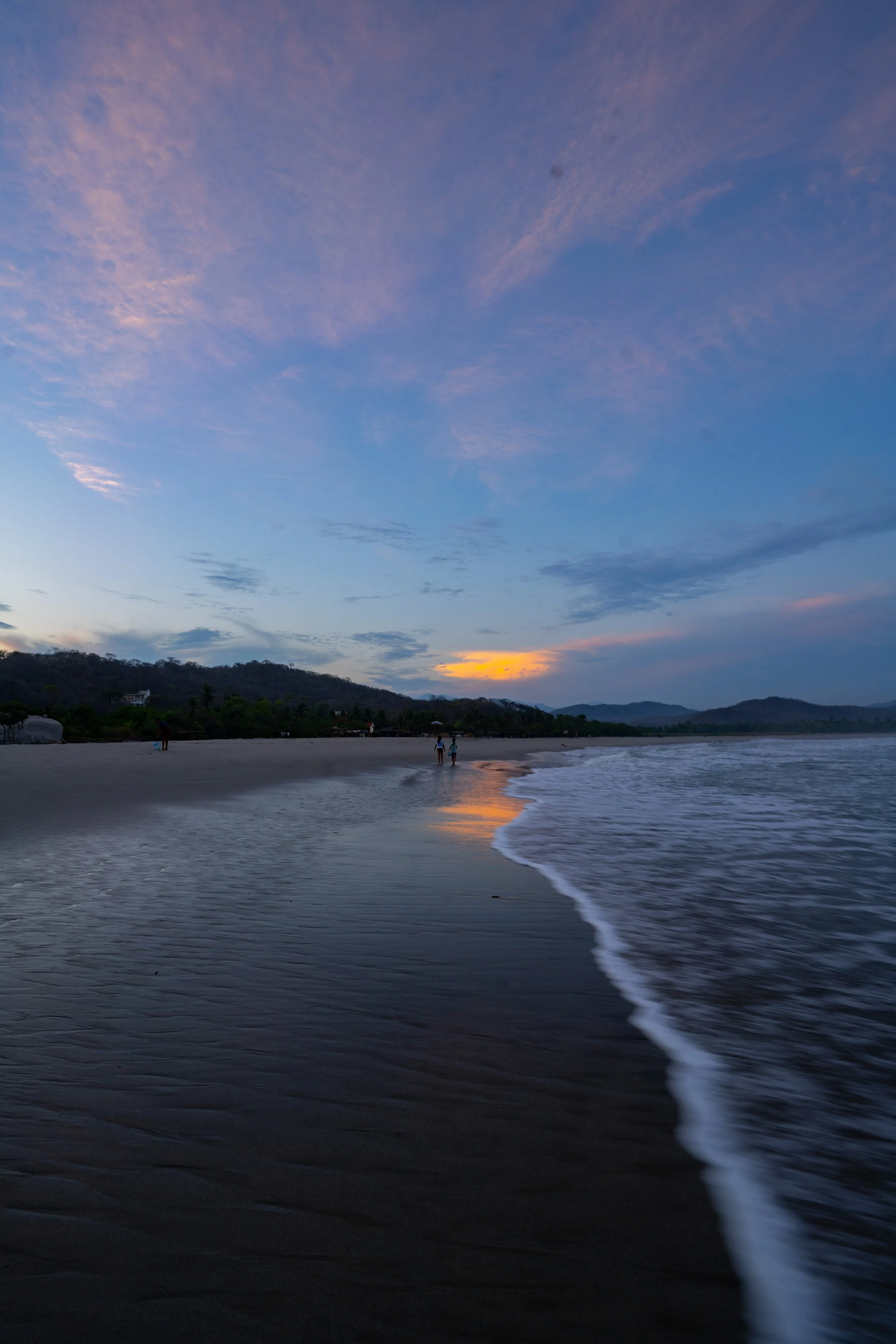 A beach at sunset with a few people walking along the shore, calm waves, and a mountain range in the background under a partly cloudy sky.