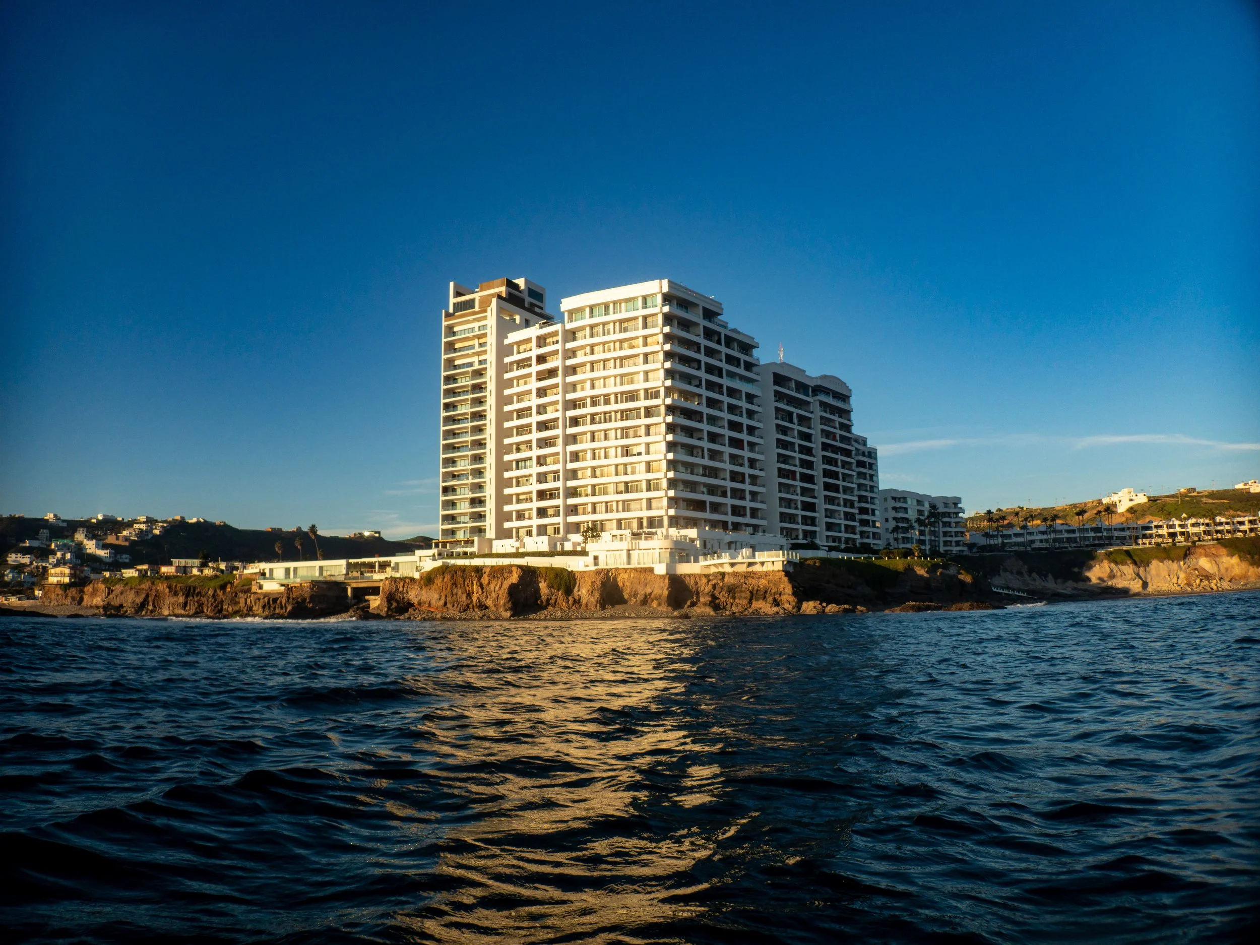A large white modern apartment building on a rocky coastline, with water in the foreground and a clear blue sky in the background.