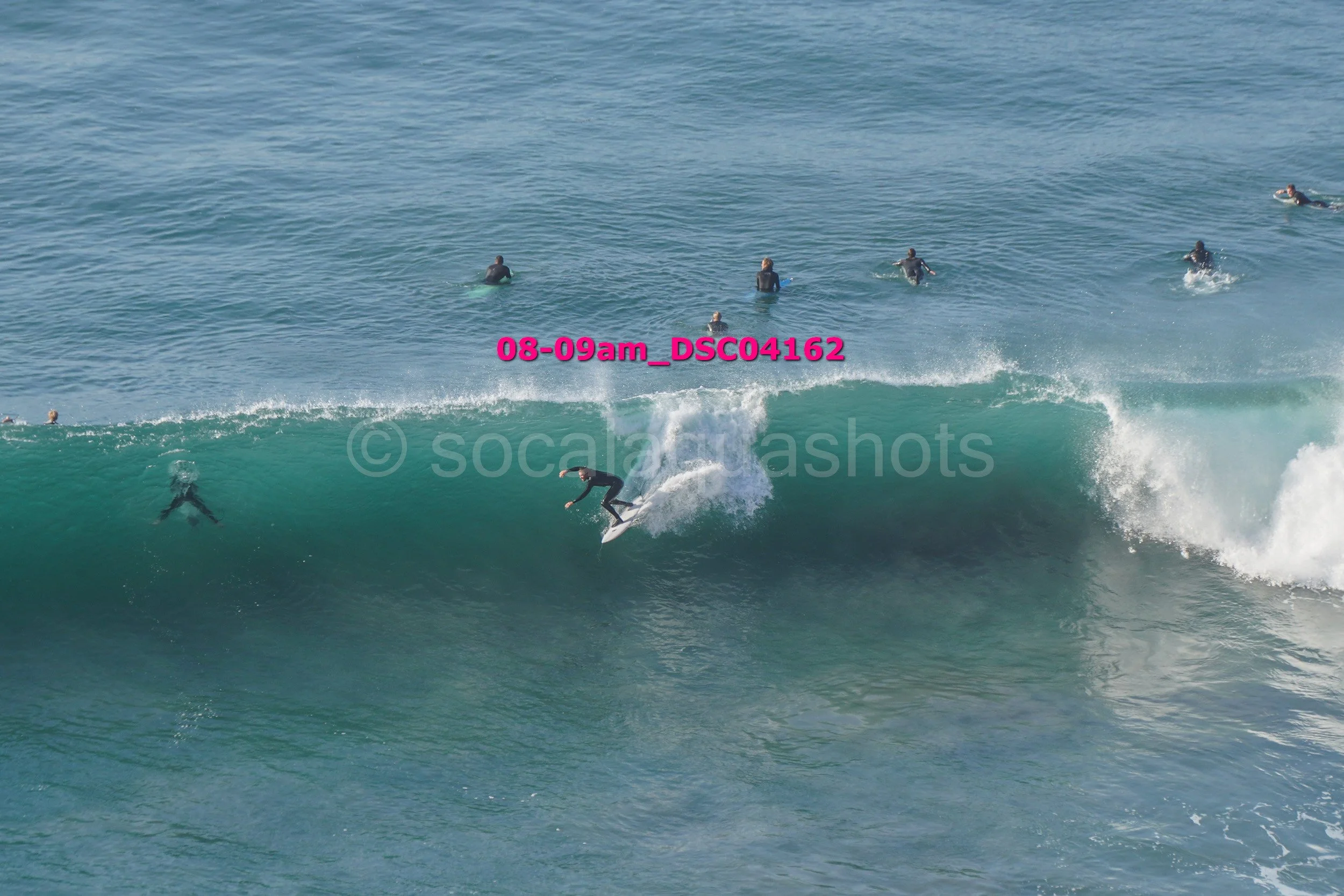 Surfer riding a large wave with several people in the water watching from behind.