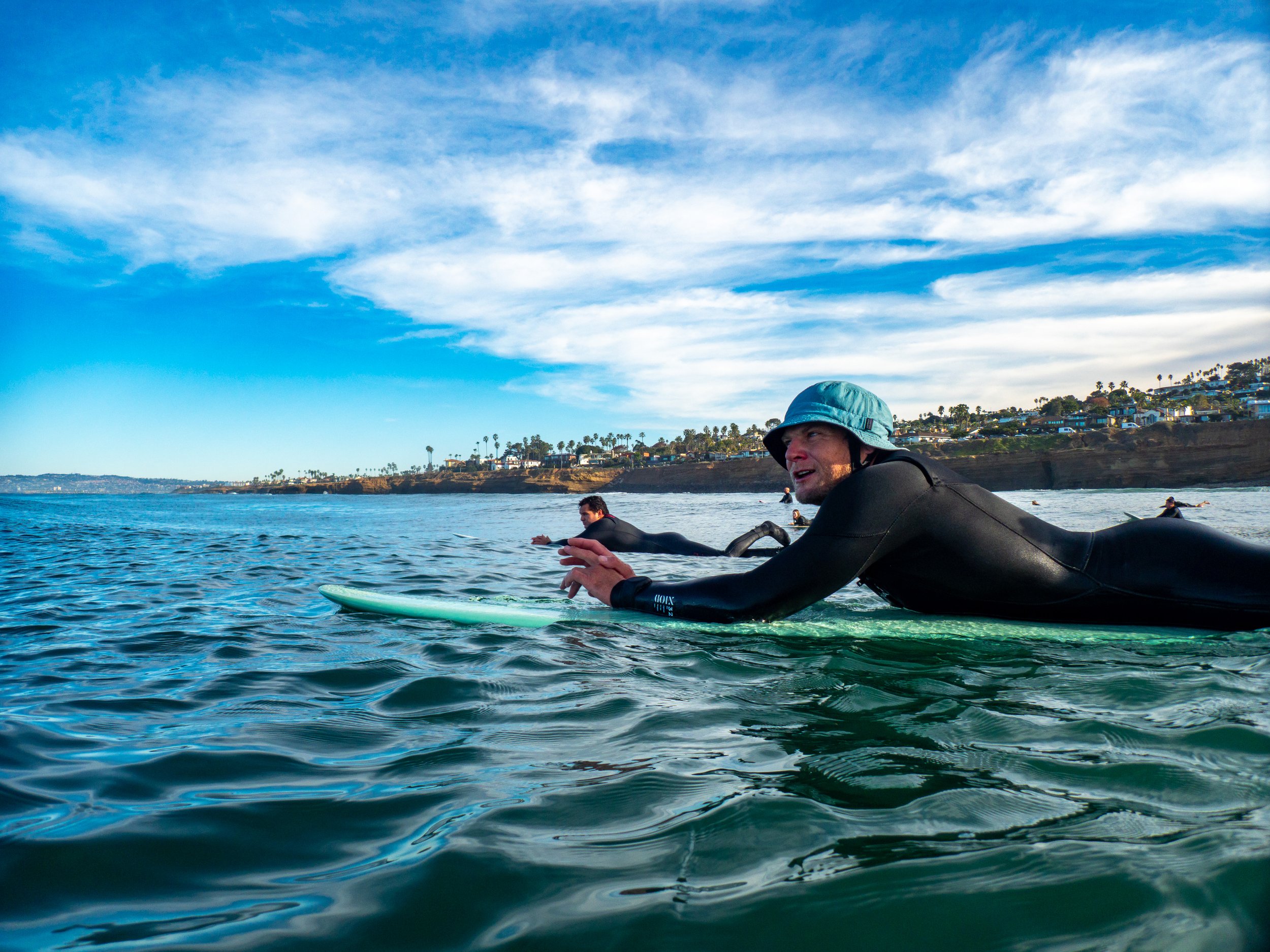 People in wetsuits surfing in the ocean near a coastline with houses and palm trees, under a sky with clouds.
