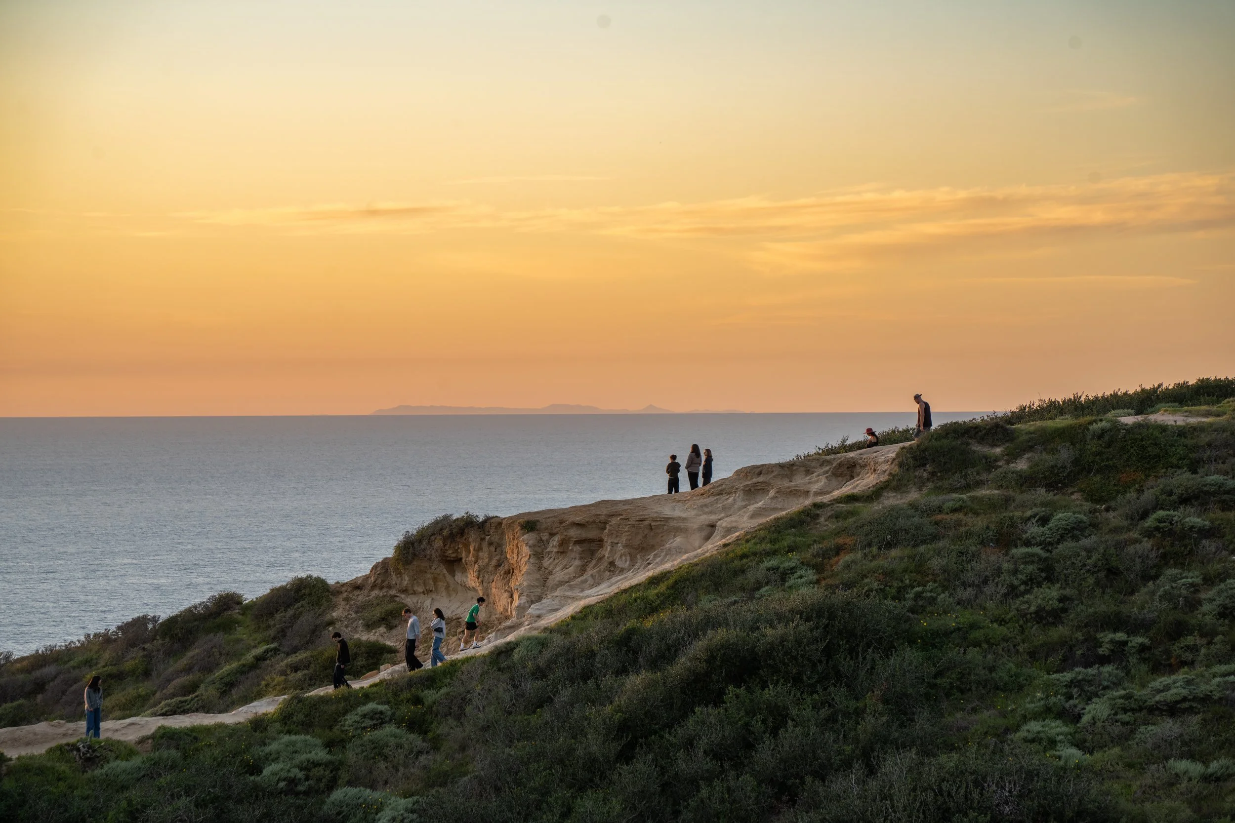 People walking along a dirt trail on a hillside overlooking the ocean at sunset, with a distant landmass visible on the horizon.