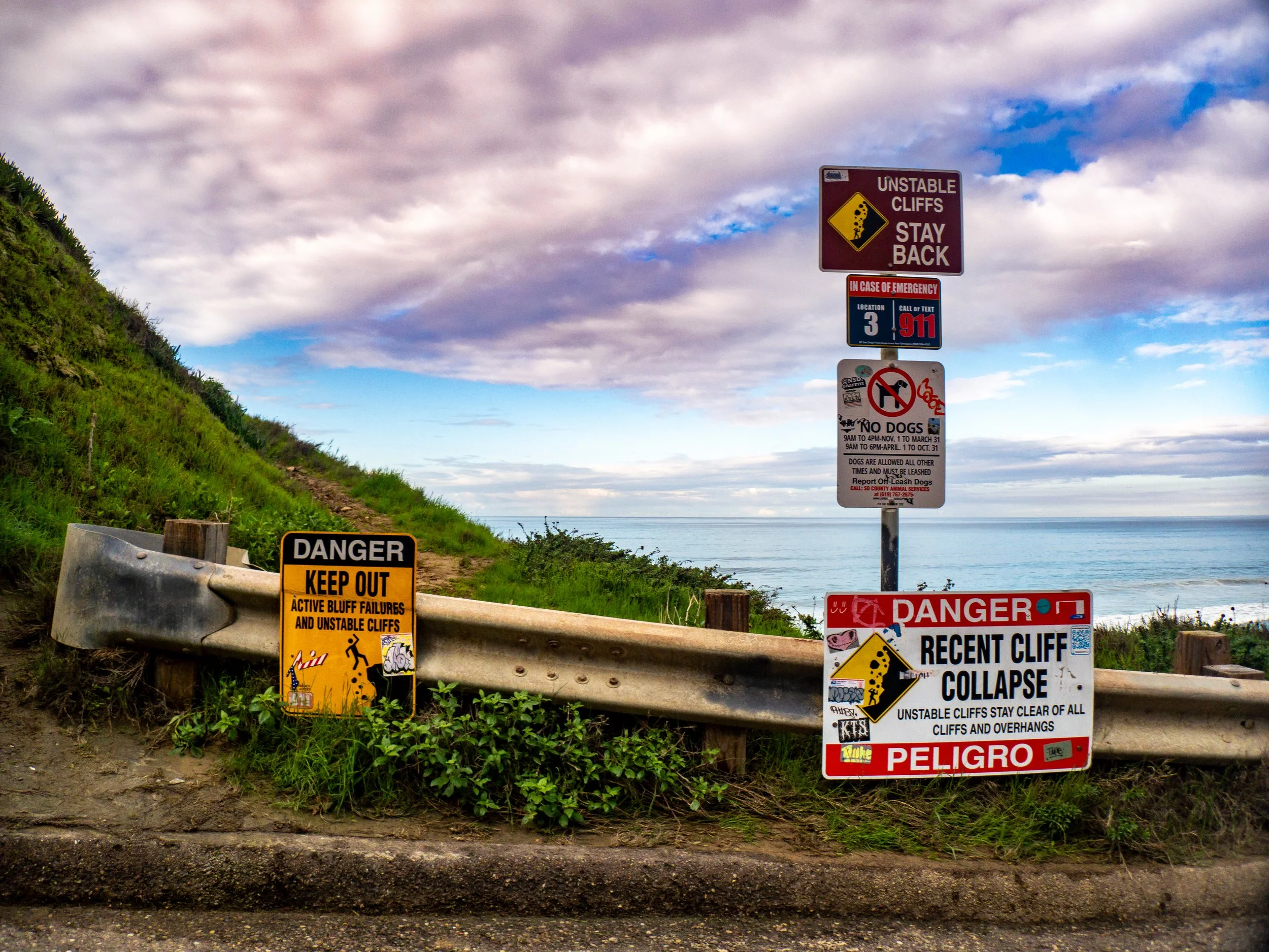 Cliffside warning signs warning of unstable cliffs and recent cliff collapse near the ocean, with a grassy hillside, cloudy sky, and ocean in the background.