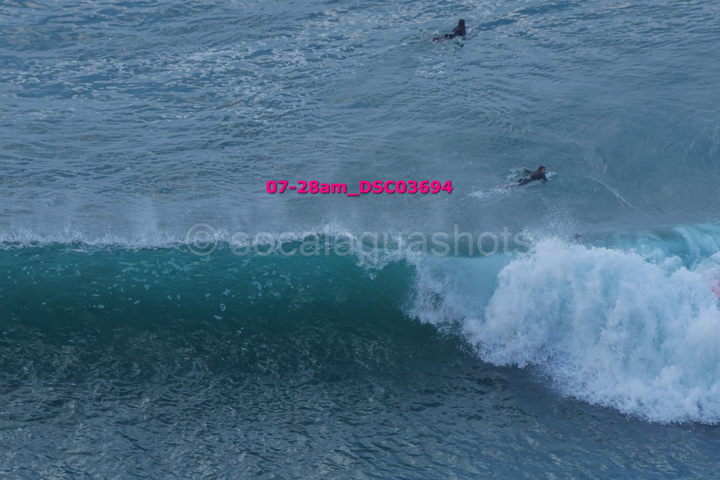 Two surfers riding a large ocean wave, with part of the wave curling over and white froth at the crest, in the open sea.