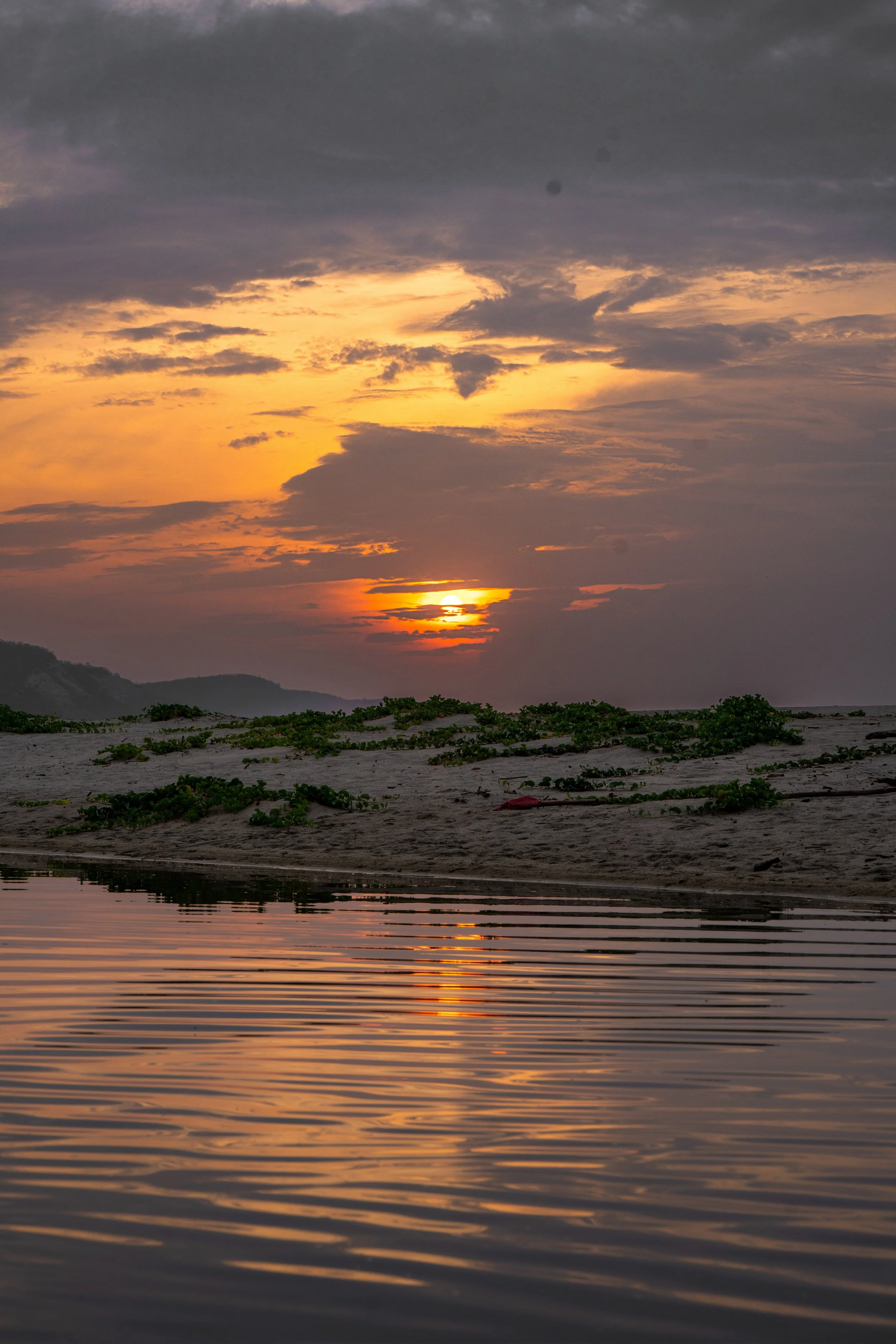 A sunset over a body of water with reflections, with a cloudy sky and a distant shoreline with hills and vegetation.