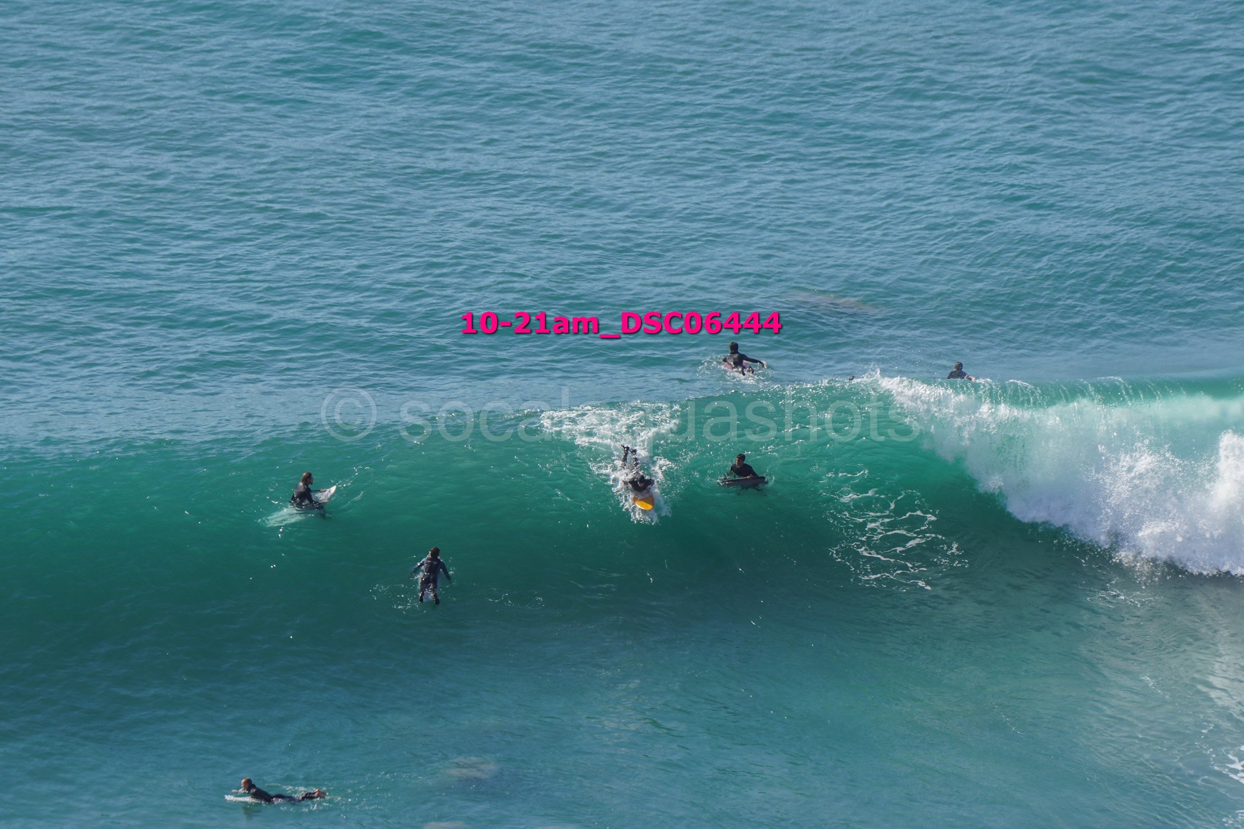Multiple surfers in wetsuits riding and paddling on a large ocean wave with clear blue water.