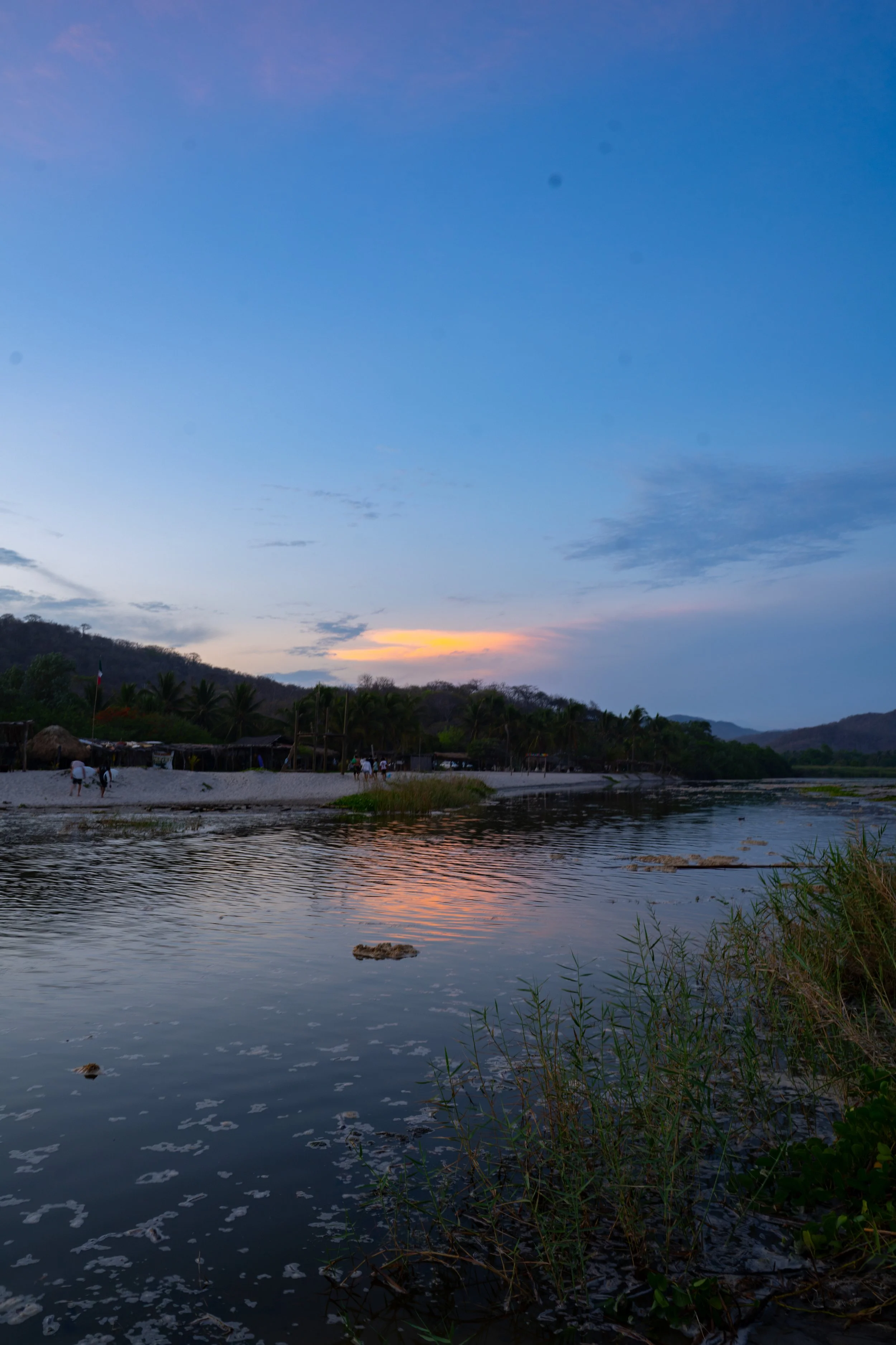 A river at sunset with a partially clouded sky and distant hills. Trees and people are visible along the riverbank.