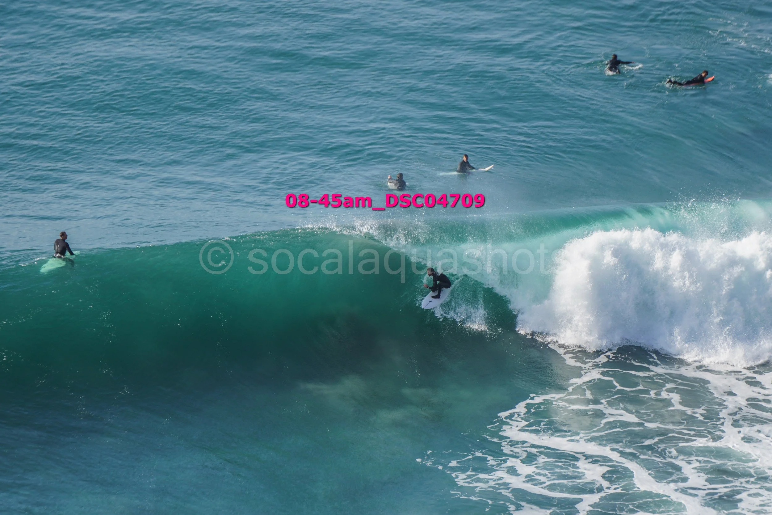 Multiple surfers in black wetsuits surfing and riding a large blue-green wave in the ocean during daytime.