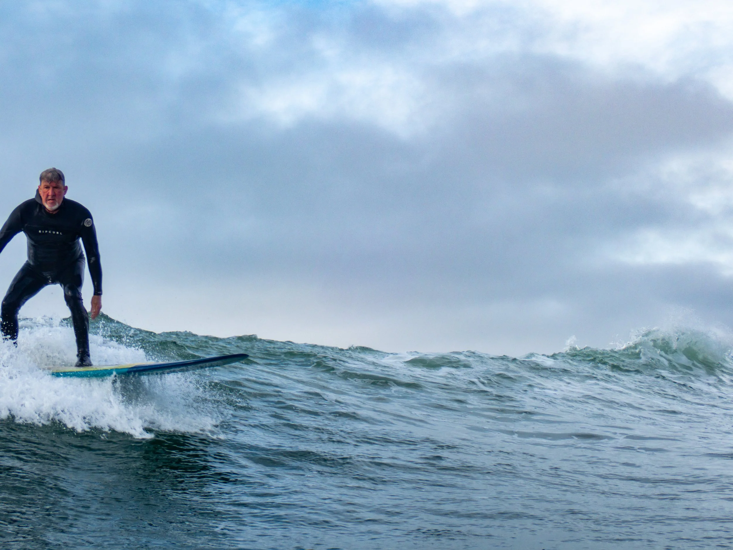 A man surfing on a wave in the ocean, wearing a wetsuit, with cloudy sky in the background.