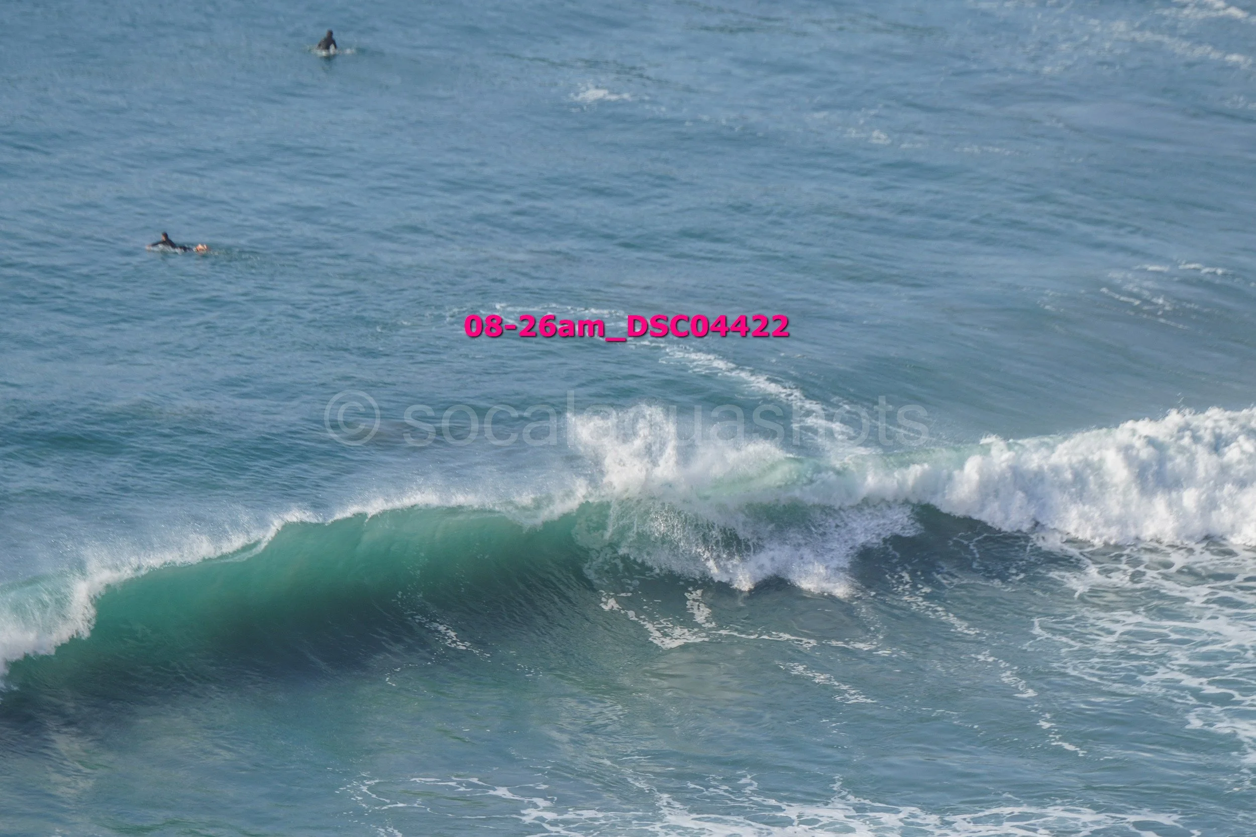 Ocean waves with three surfers in the distance, one closer to the camera, with a timestamp and file name overlaid.