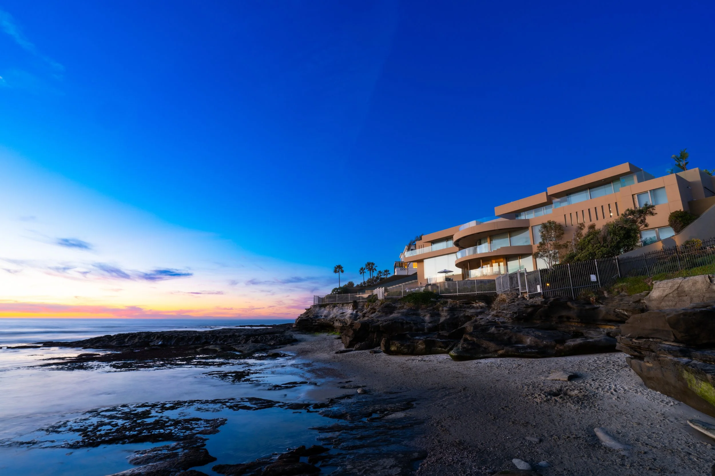 Modern beachfront house on rocky shoreline during sunset with colorful sky and palm trees in background.