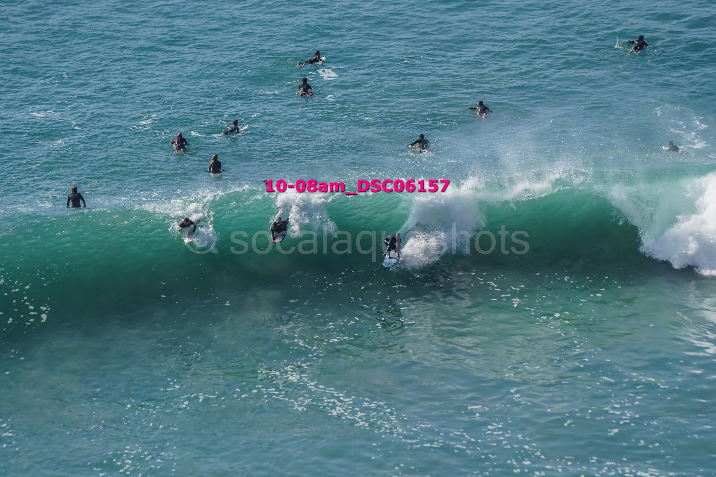 Multiple surfers riding waves and waiting in the ocean, with some surfing the wave and others floating or paddling.