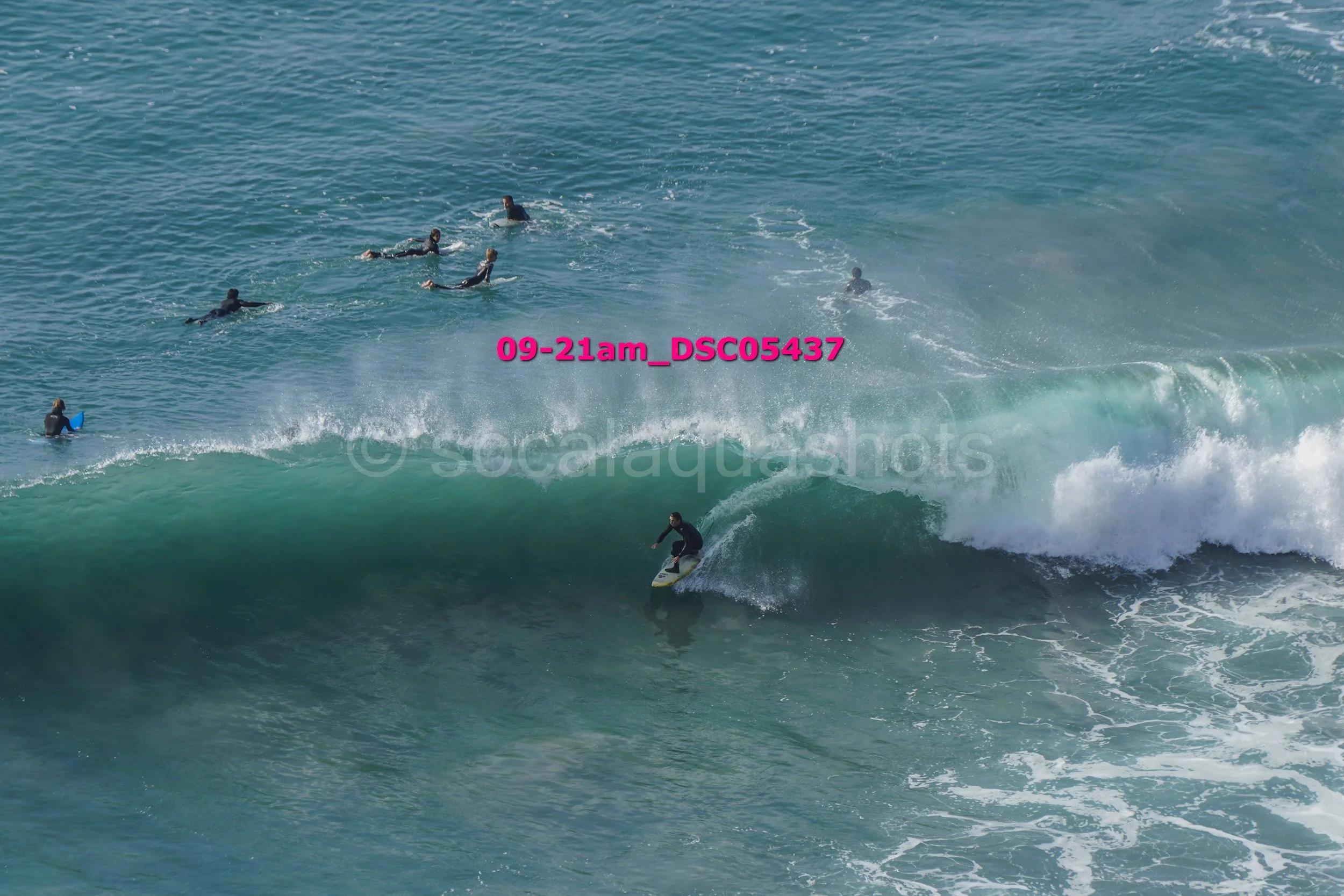A surfer riding a large wave while several people swim and surf in the background.