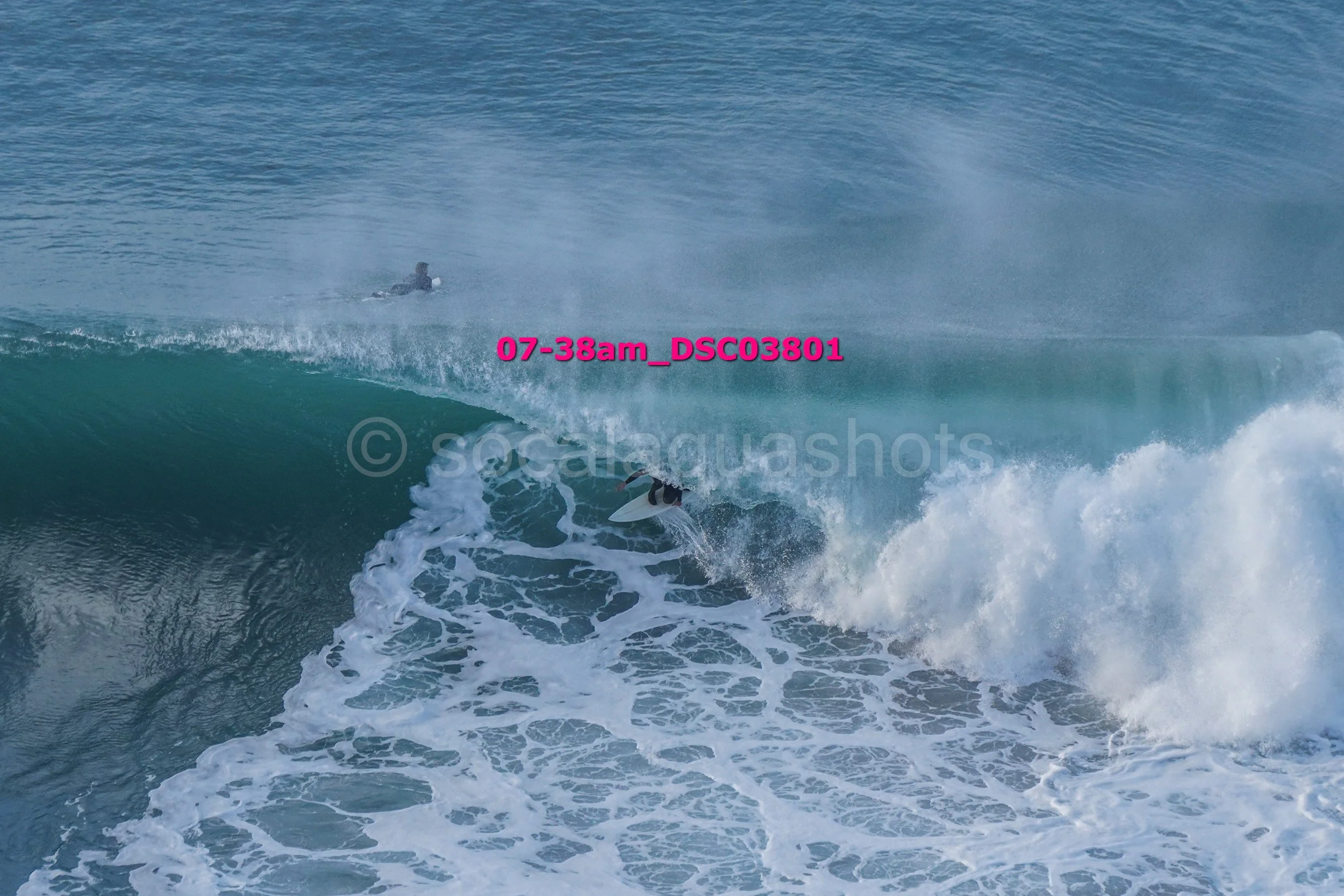 A surfer riding a large wave in the ocean with another person swimming in the background.