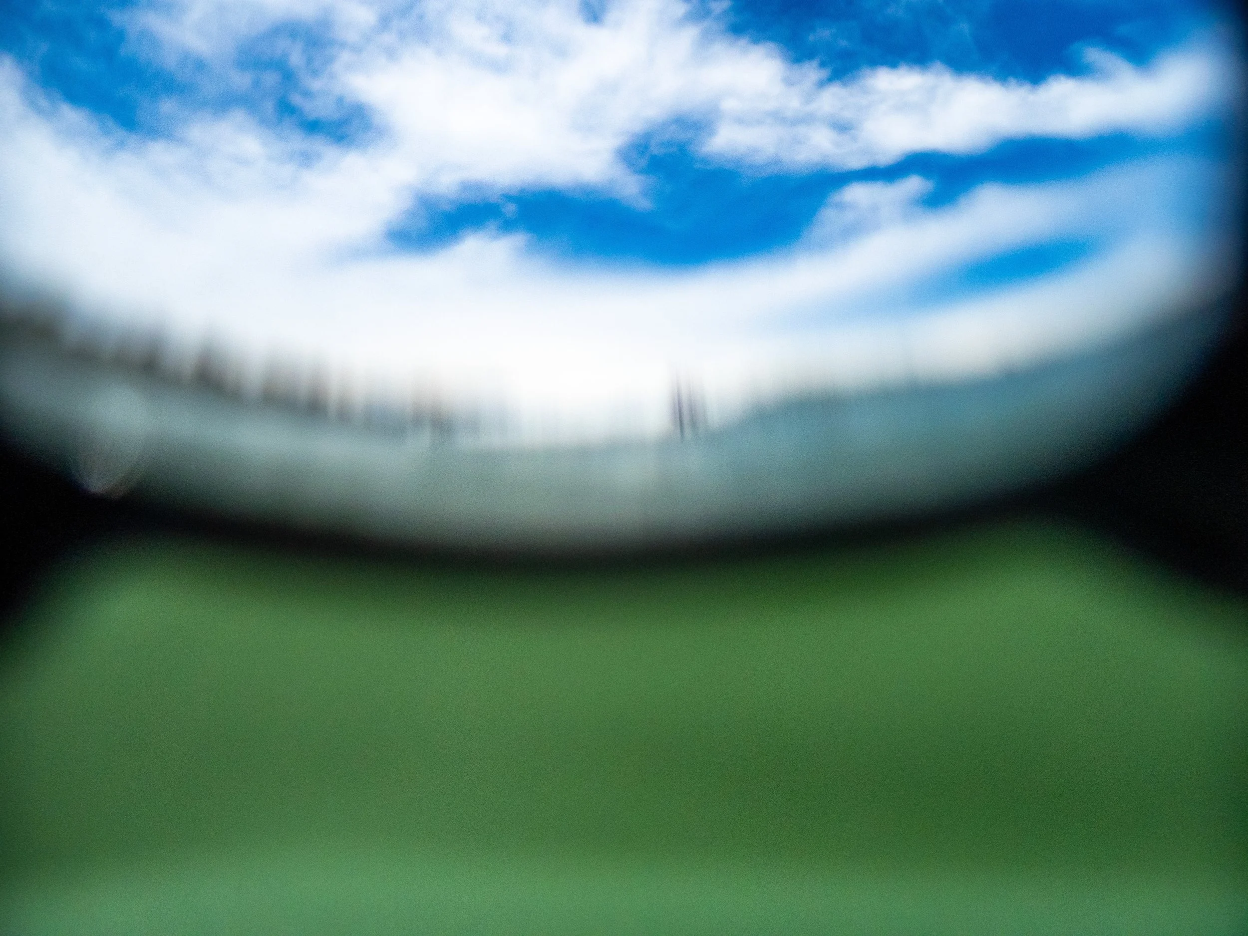 Blurred image showing a patch of blue sky with white clouds and some green below, viewed through an out-of-focus round object.