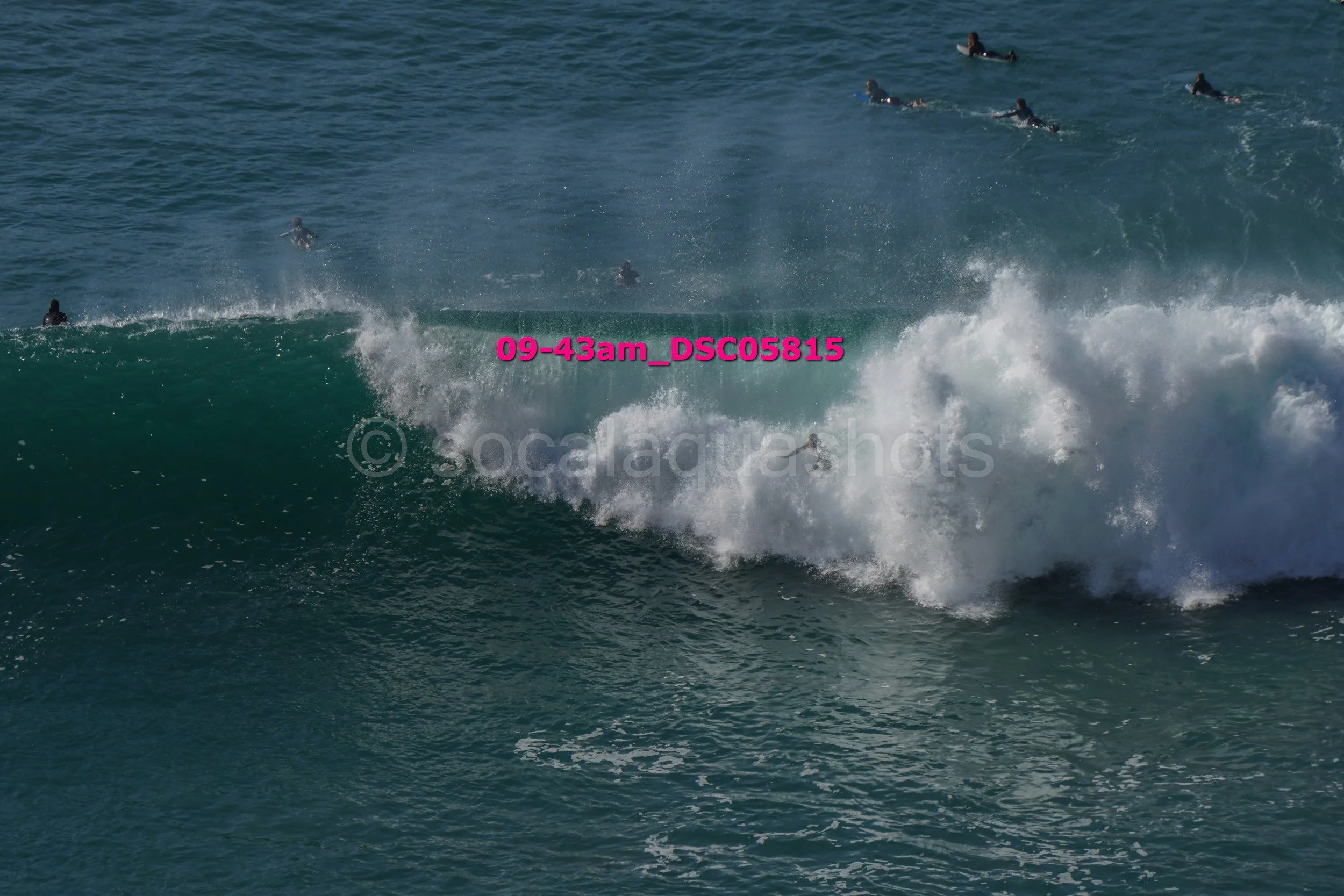 Multiple surfers in the ocean, with one large breaking wave in the foreground and others further in the distance.