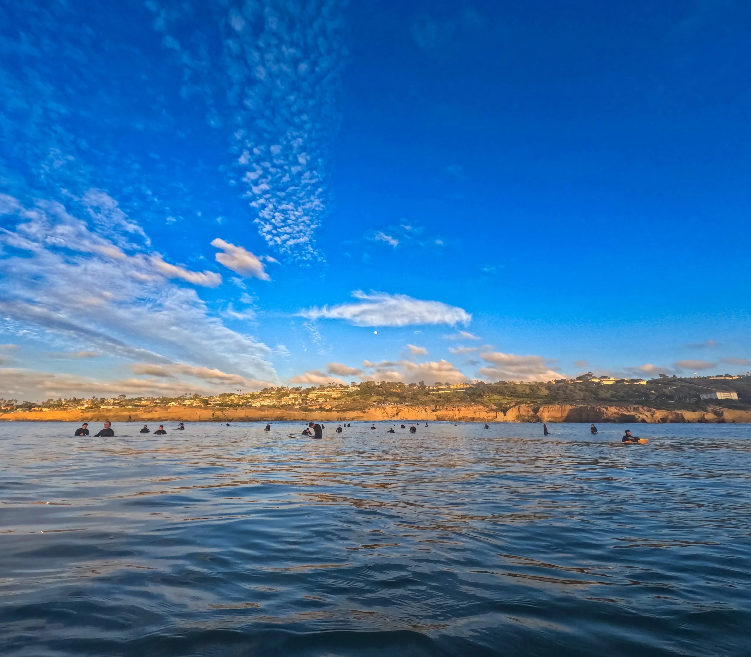 People surfing and swimming in the ocean near a coastline with hills and houses under a blue sky with clouds.