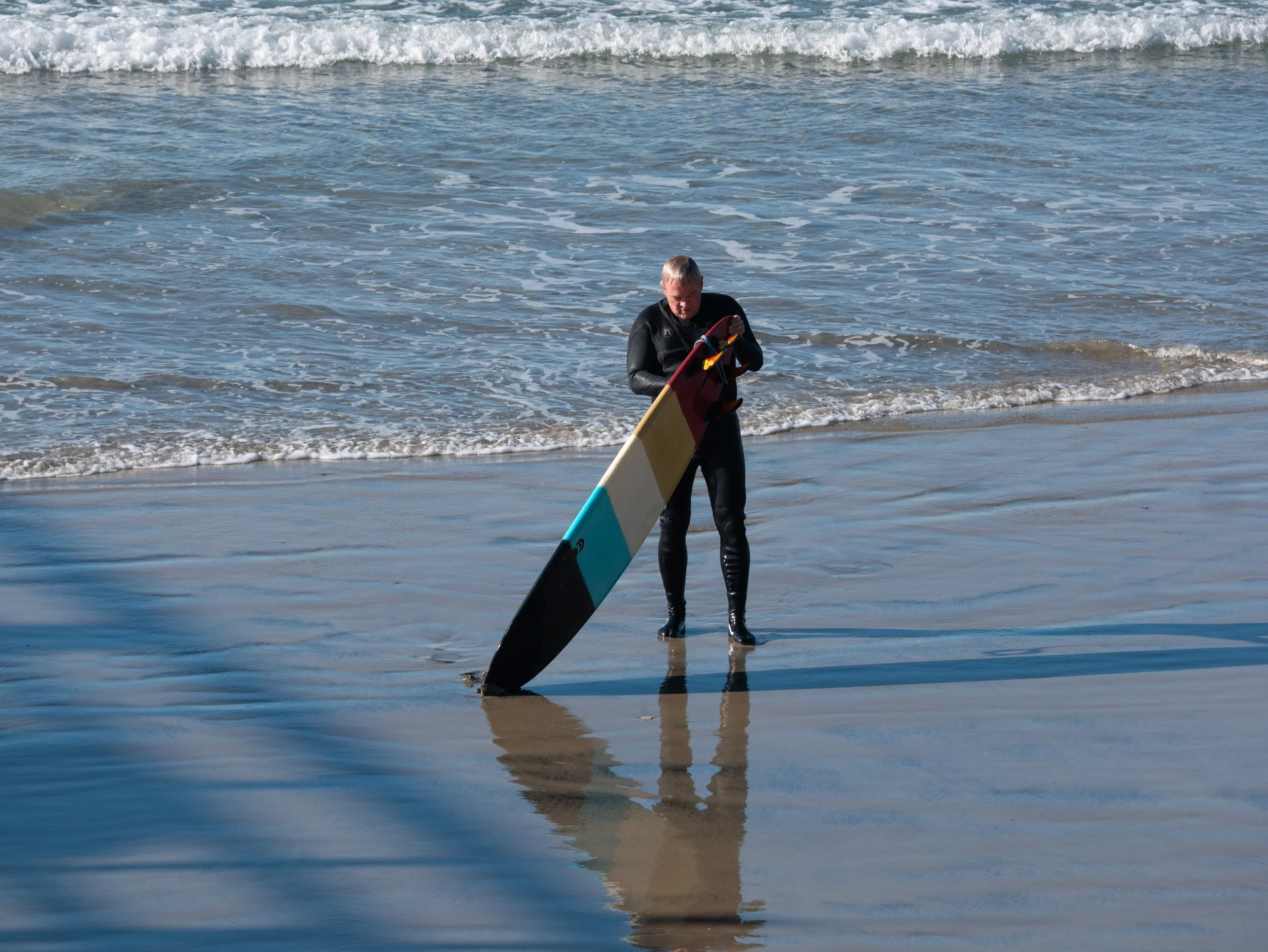 Person in a wetsuit standing in shallow water on a sandy beach, holding a colorful stand-up paddleboard.