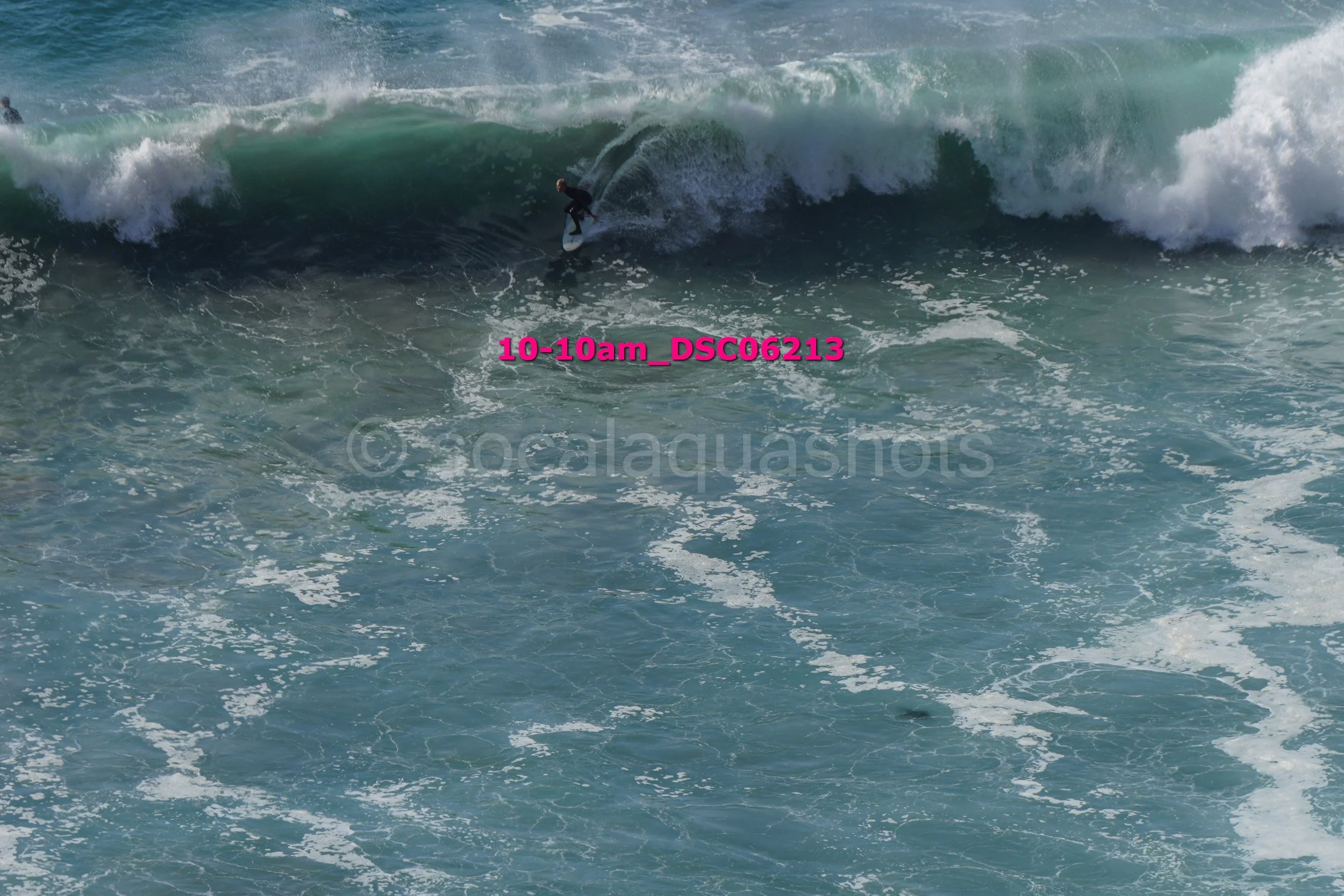 A person surfing on a large ocean wave with white foam, water in shades of blue and green, during daytime.