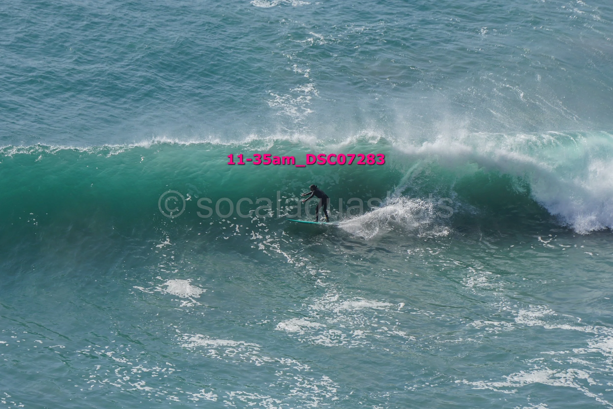 A surfer riding a large wave in the ocean with visible white foam and spray, wearing black wetsuit, with the wave cresting behind him.