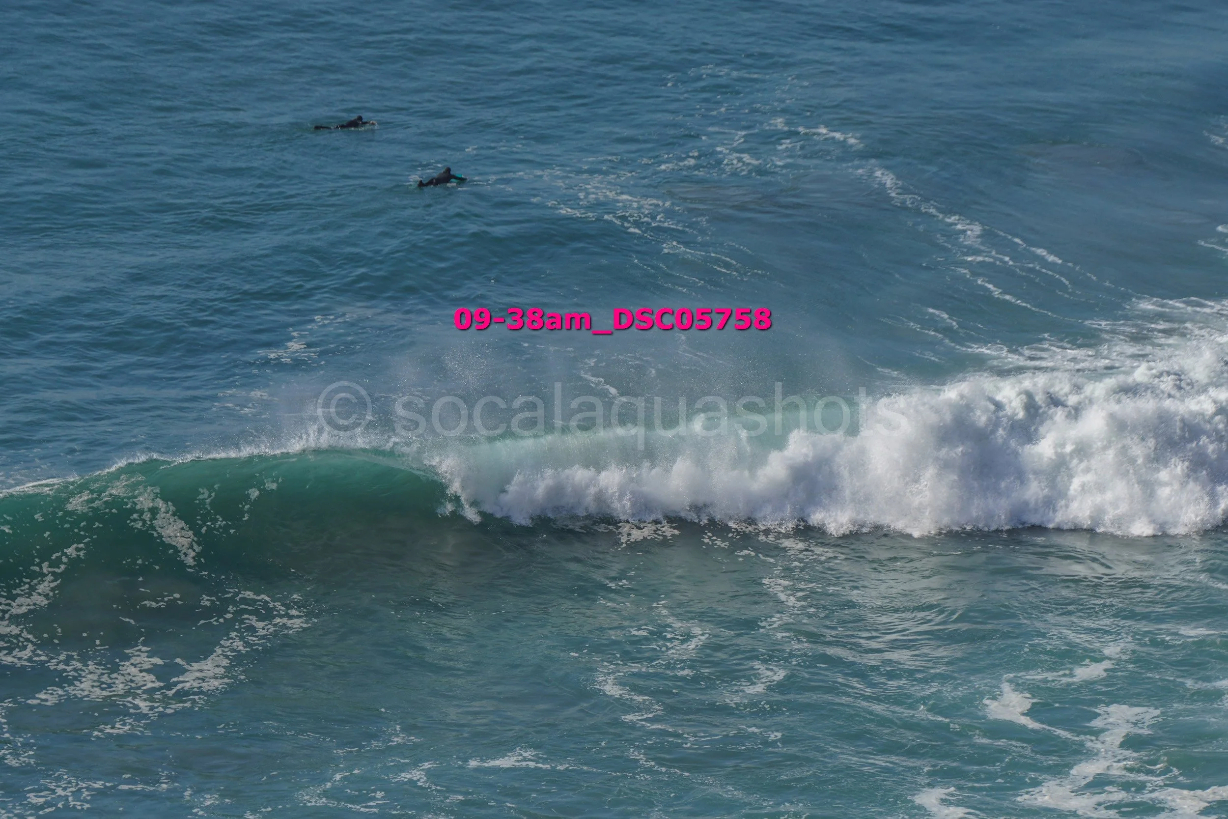 A scene at the ocean with waves crashing and two surfers in the distance, one of them riding a wave while the other floats on their surfboard.