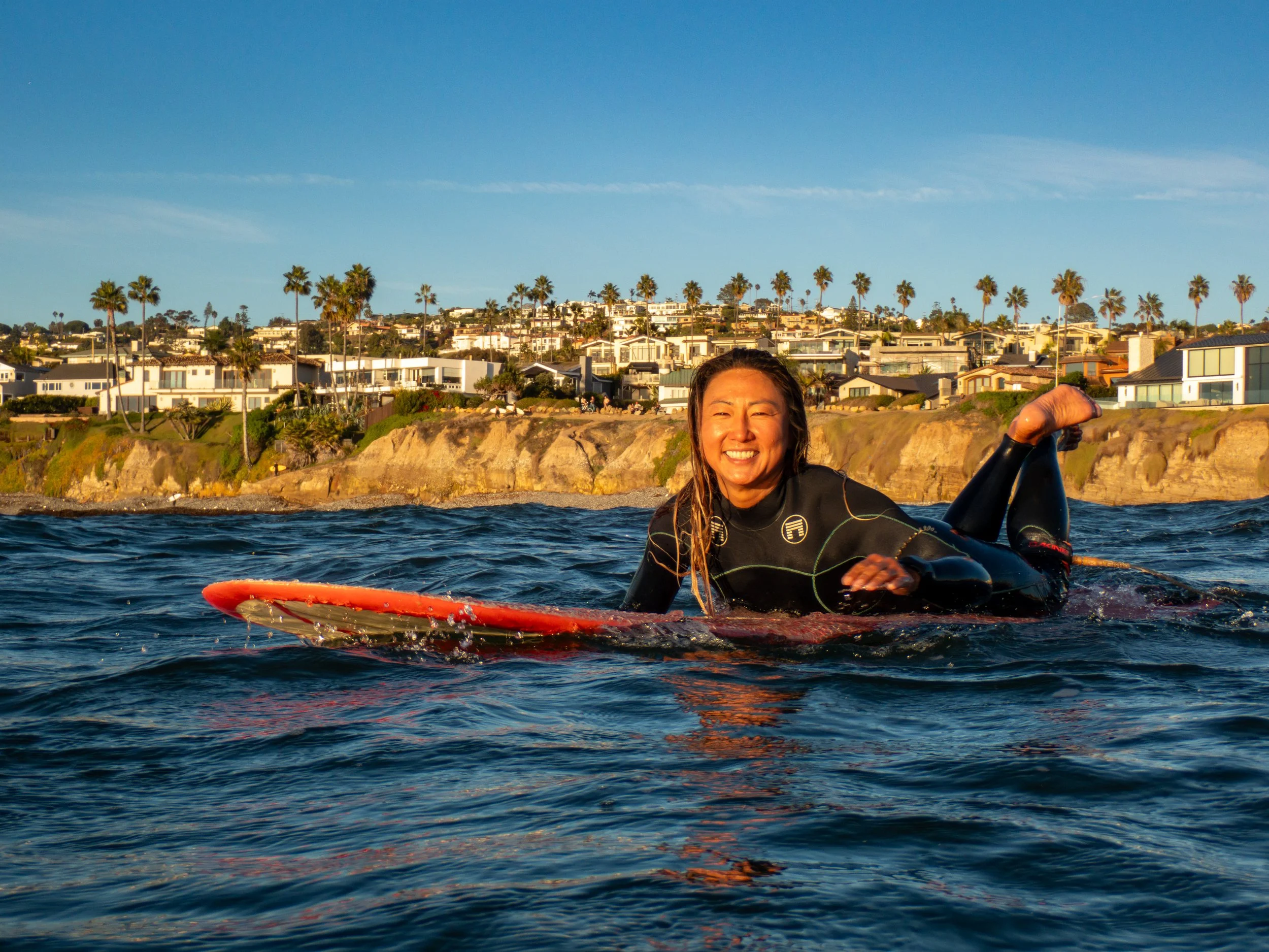 Woman smiling and lying on her stomach on a surfboard in the water with a coastal neighborhood and palm trees in the background during sunset.